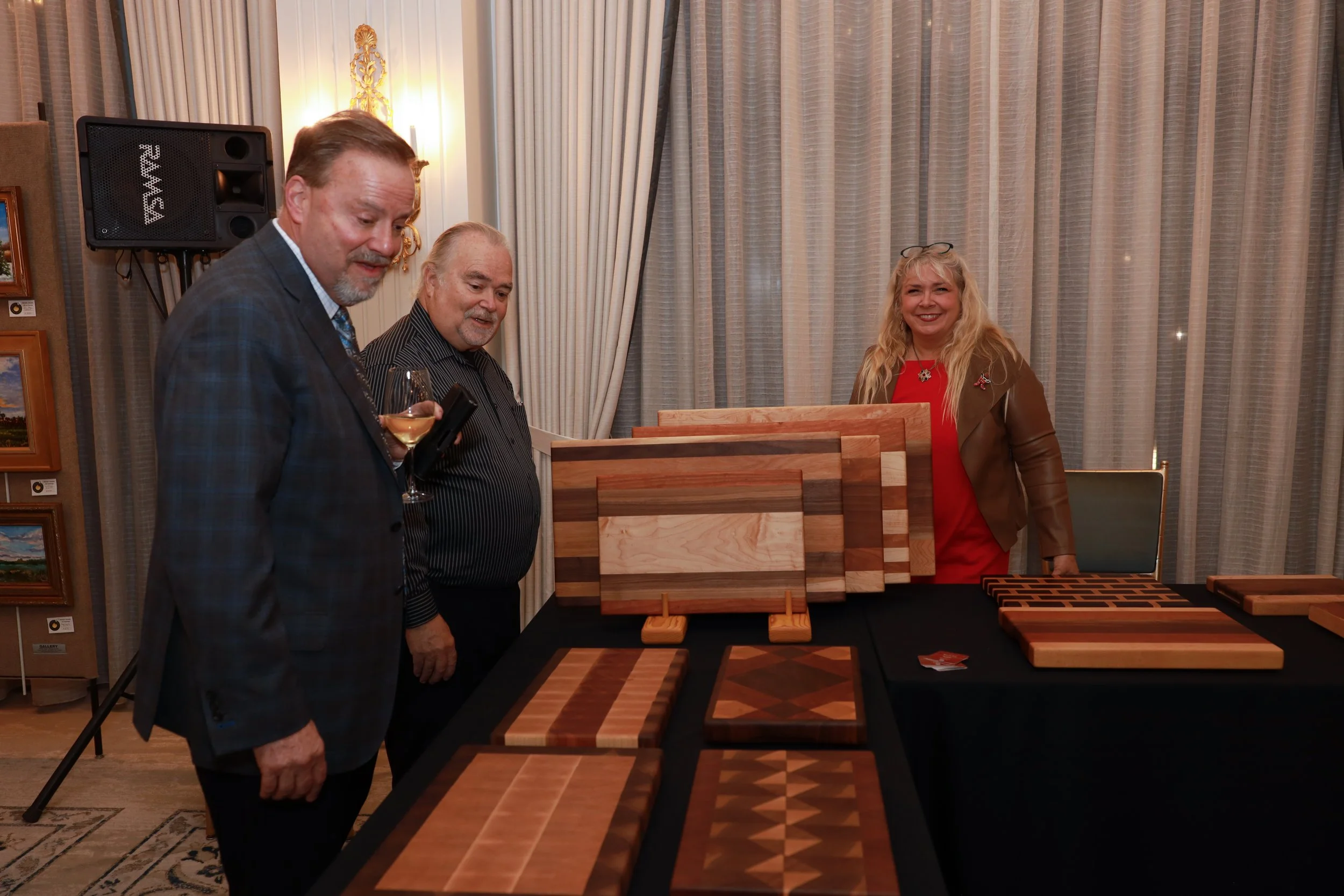 Three people, two men and one woman, at a furniture exhibition, examining wooden chessboards on a table. Behind them are large curtains and a wall with framed paintings, event lighting, and a speaker.