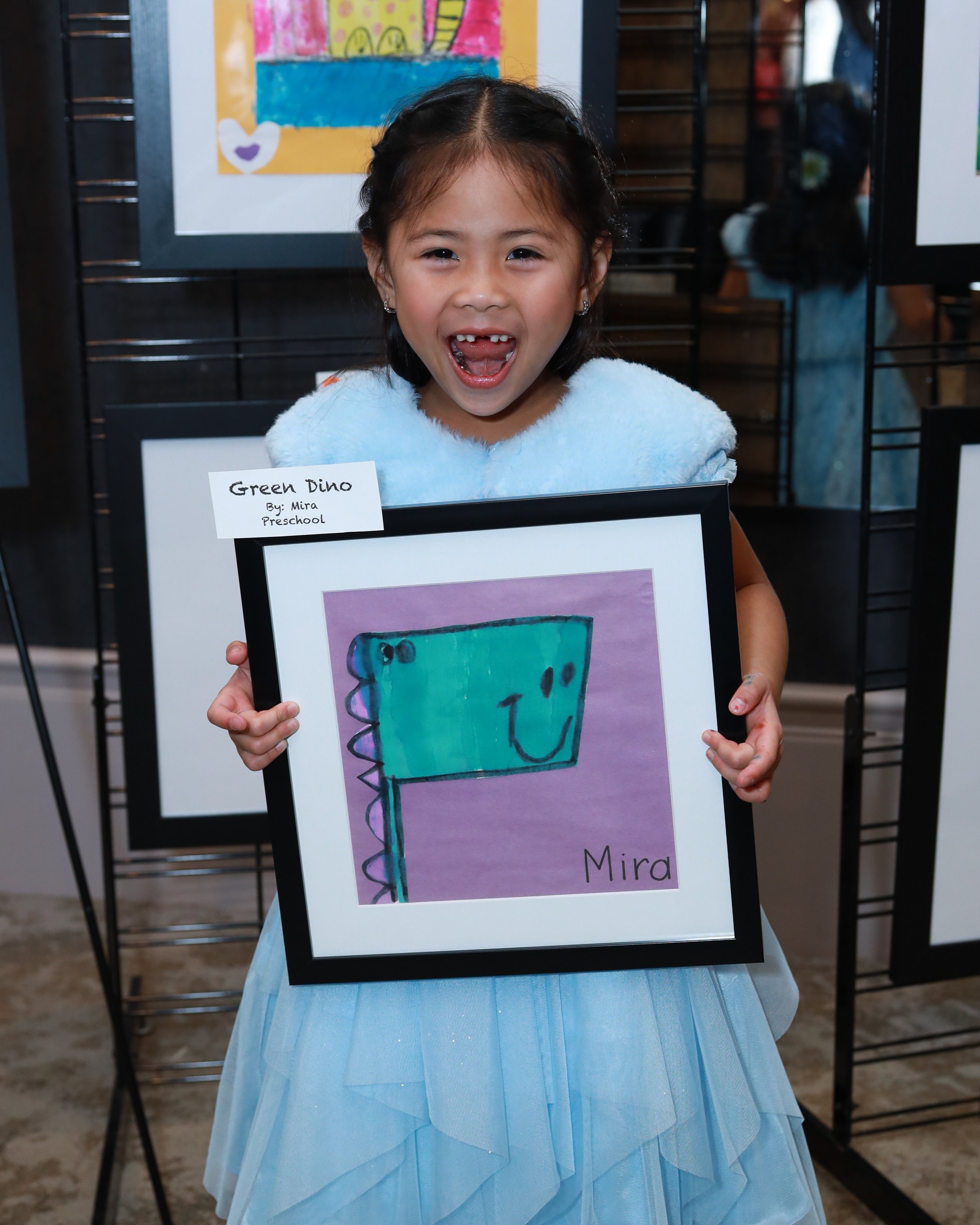 A young girl holding a framed artwork of a colorful dinosaur. The girl is smiling with an open mouth showing missing teeth. She is wearing a light blue dress. The background shows some artwork displayed on a black stand.
