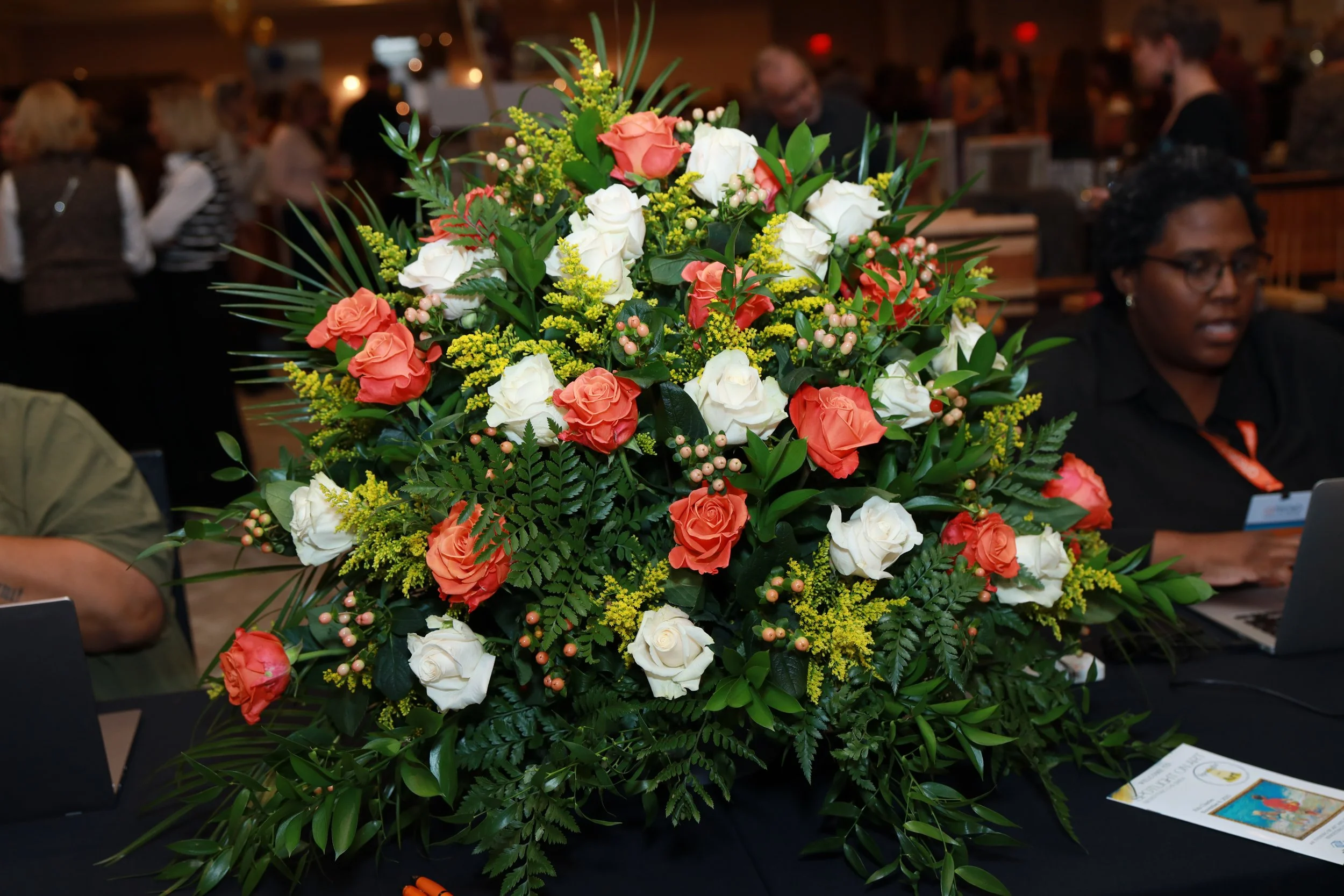 A large bouquet of white and coral roses with green foliage and small pink berries on a table at an indoor event with people in the background.