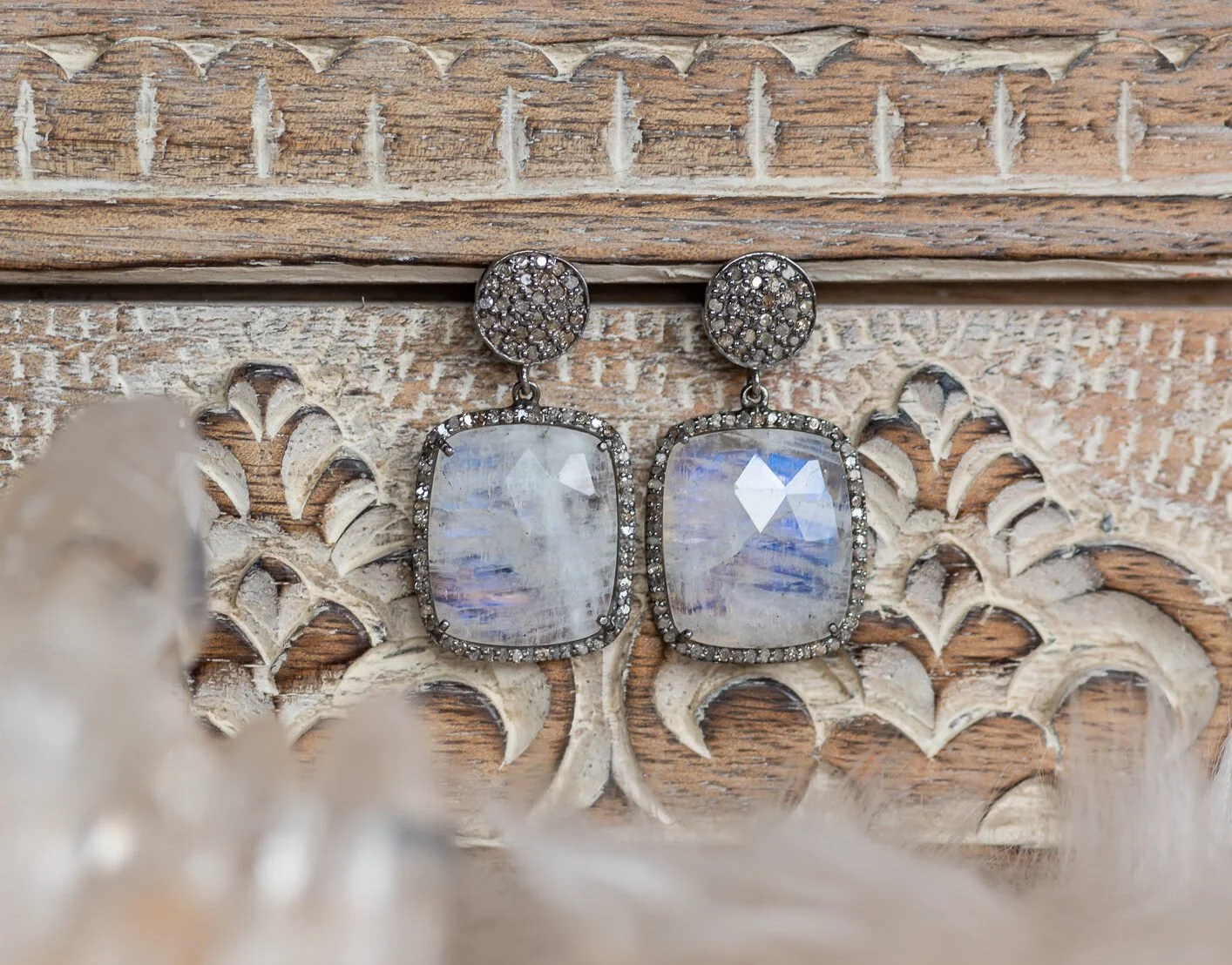 Pair of earrings with large rectangular moonstone gemstones surrounded by small rhinestones, hanging from round rhinestone-studded posts, resting on a carved wooden surface.