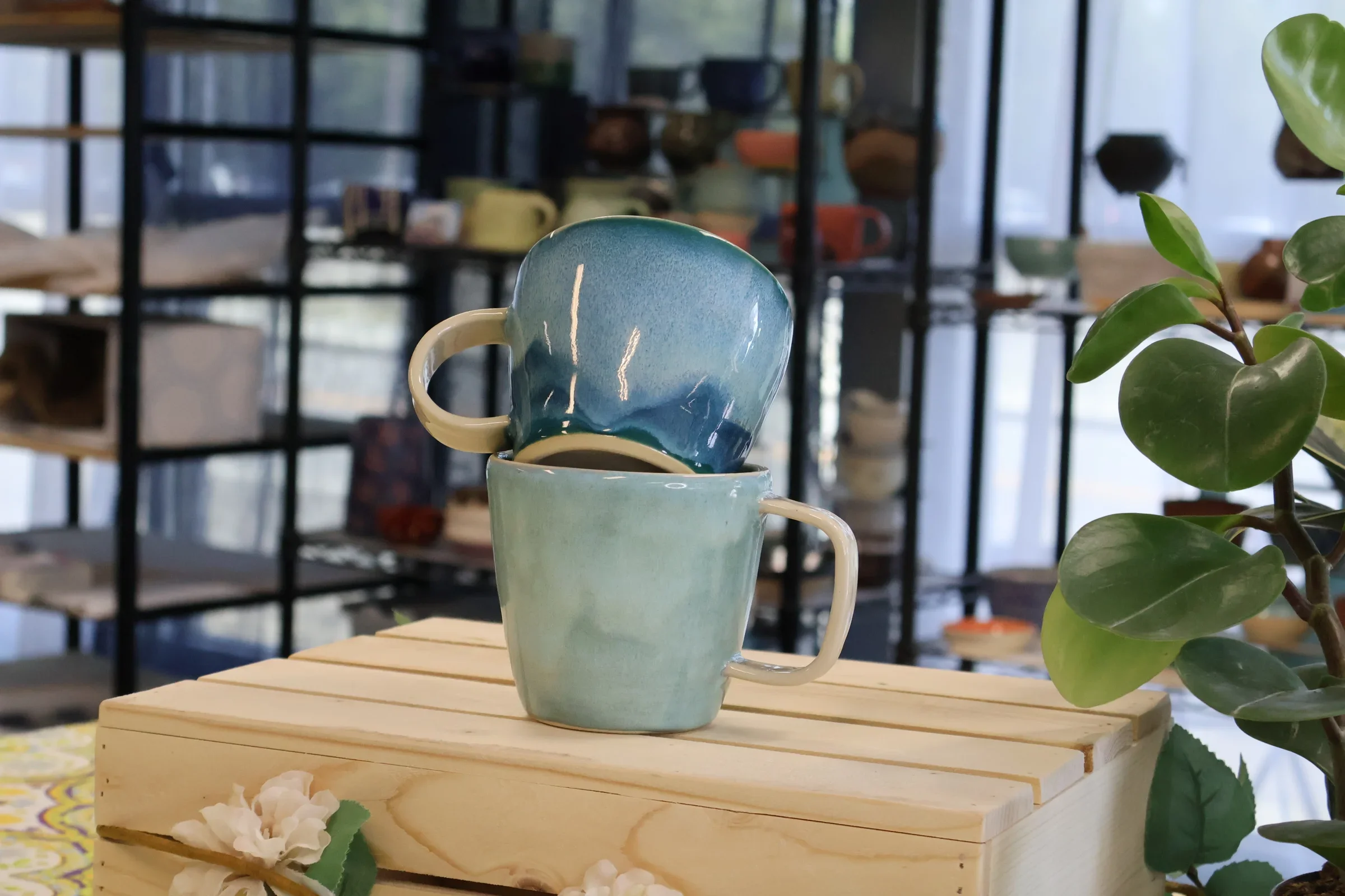 Stacked blue and cream ceramic mugs on a wooden surface with a potted plant nearby, against the backdrop of shelves with various pottery and cups.