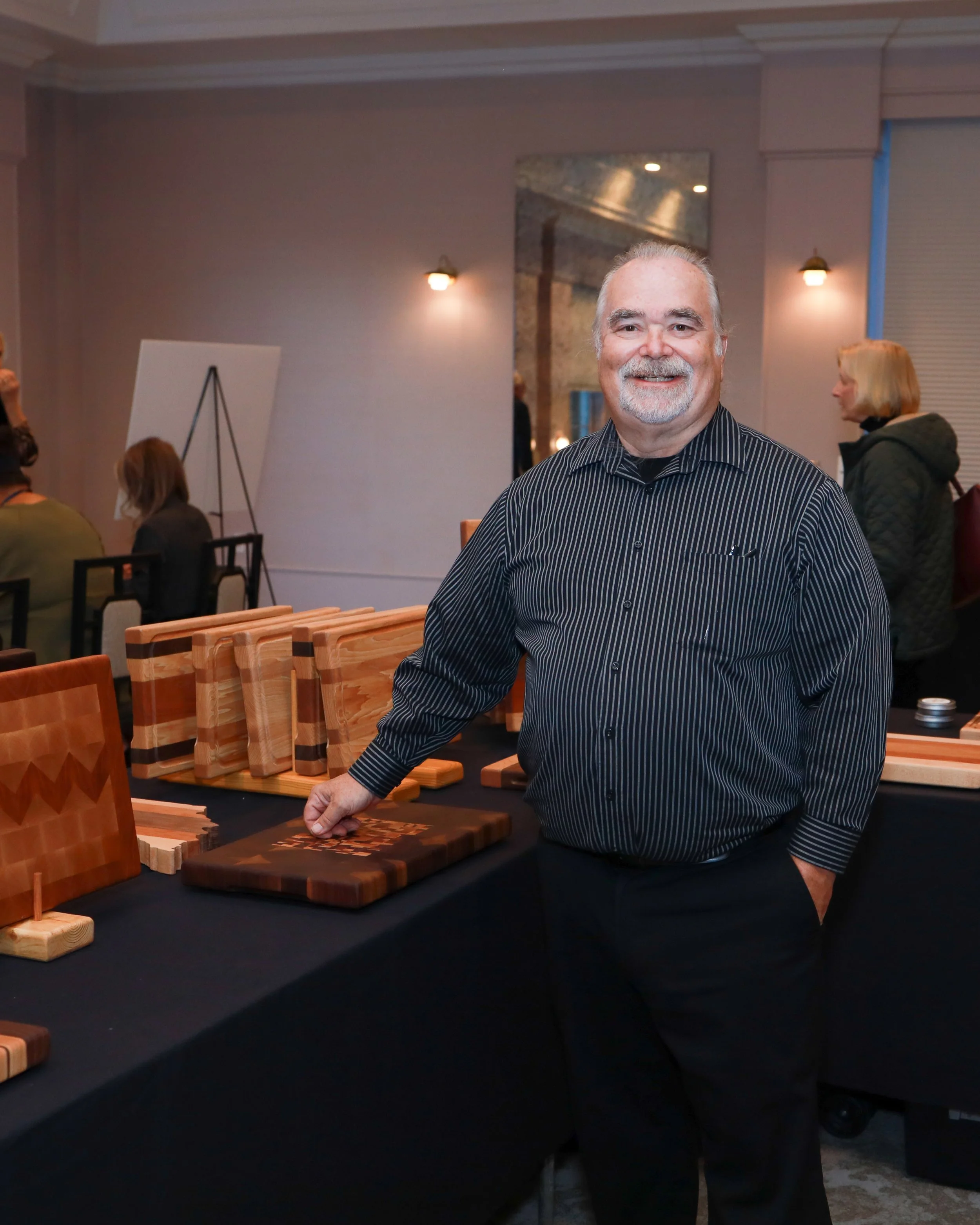 A smiling man in a black striped shirt standing at a table displaying wooden chess boards at an indoor event.