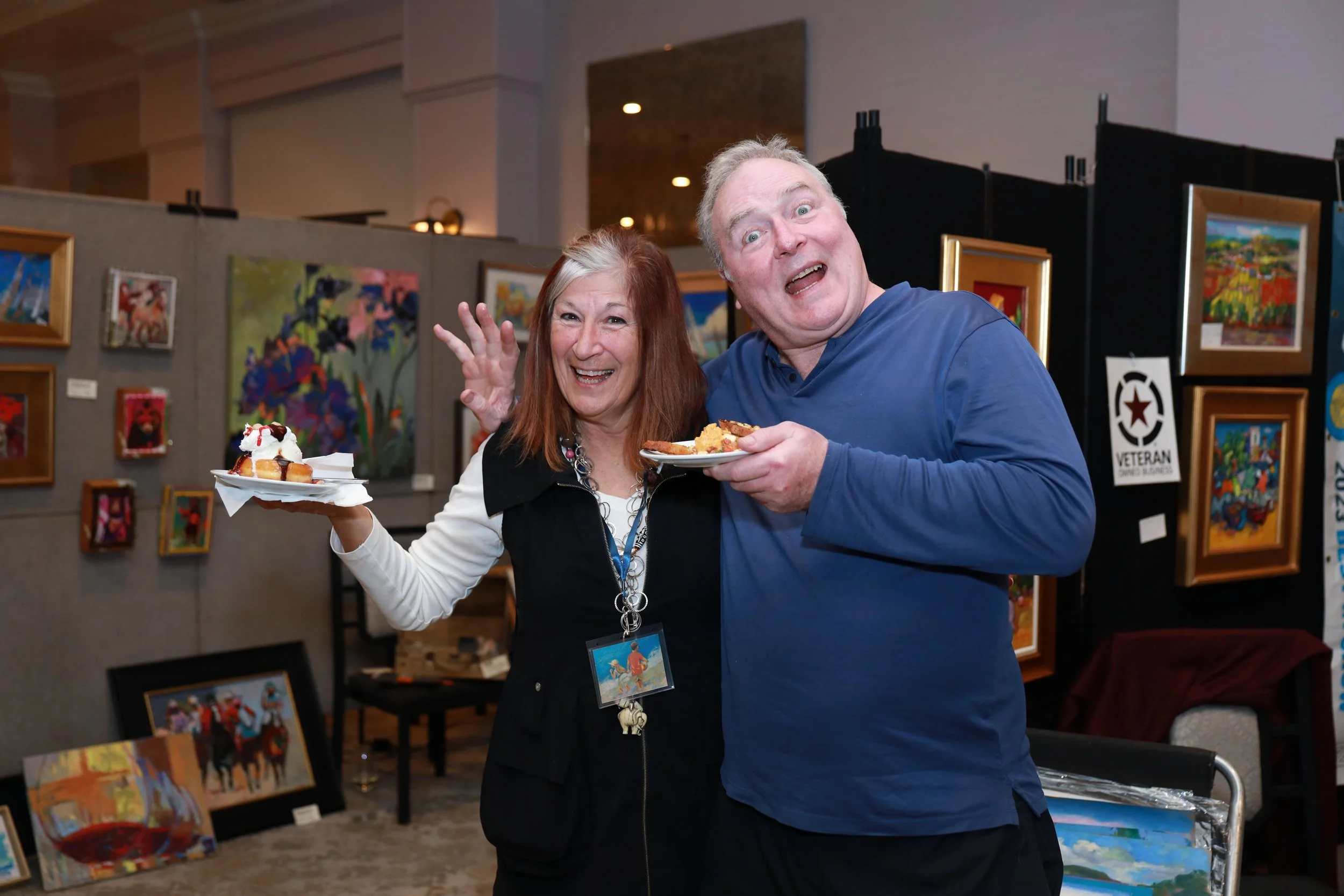A man and a woman at an art exhibition, holding plates of food, smiling at the camera, surrounded by colorful paintings displayed on the walls.