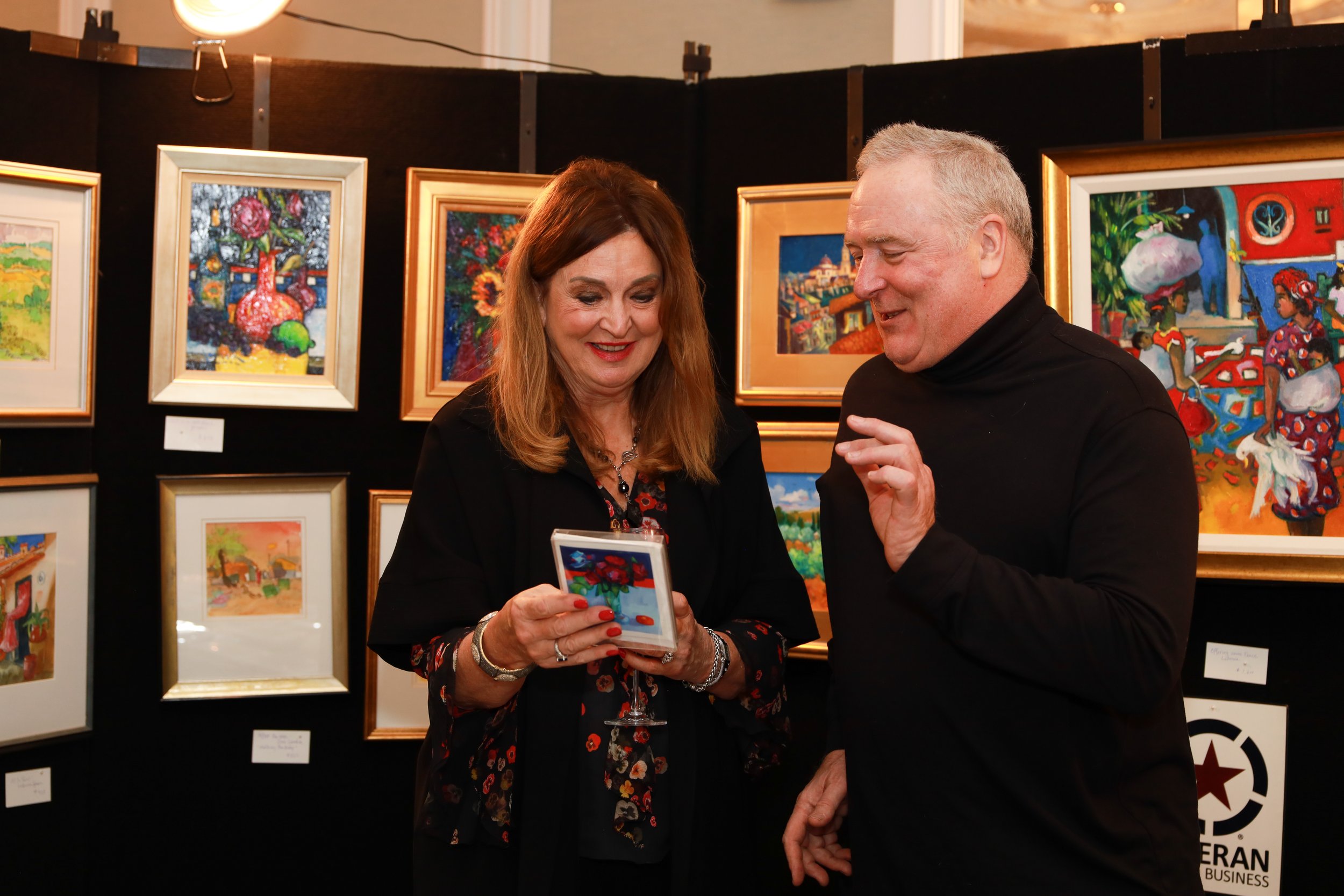 A woman and a man are looking at a small colorful painting together at an art gallery. The gallery has framed paintings hanging on the wall behind them.