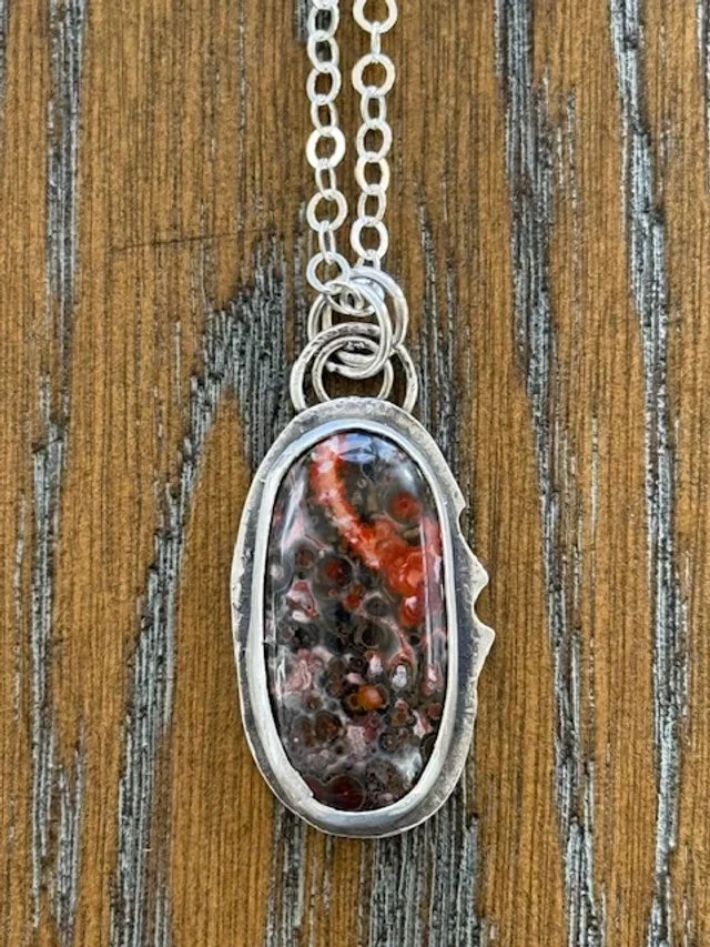 A necklace with an oval pendant featuring a marine-themed design, including small red, orange, and black coral pieces encased in clear resin, hanging from a silver chain on a wooden surface.