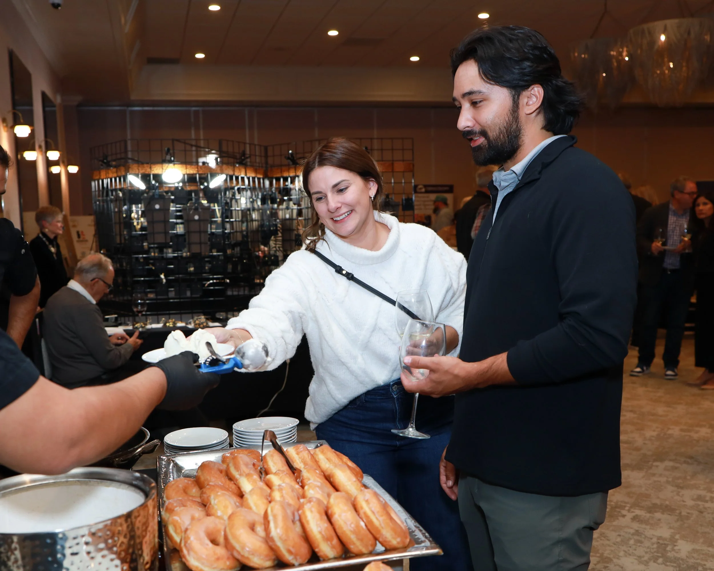 A woman and a man enjoying food at a buffet table with bread and pastries, indoors at a social gathering or event.