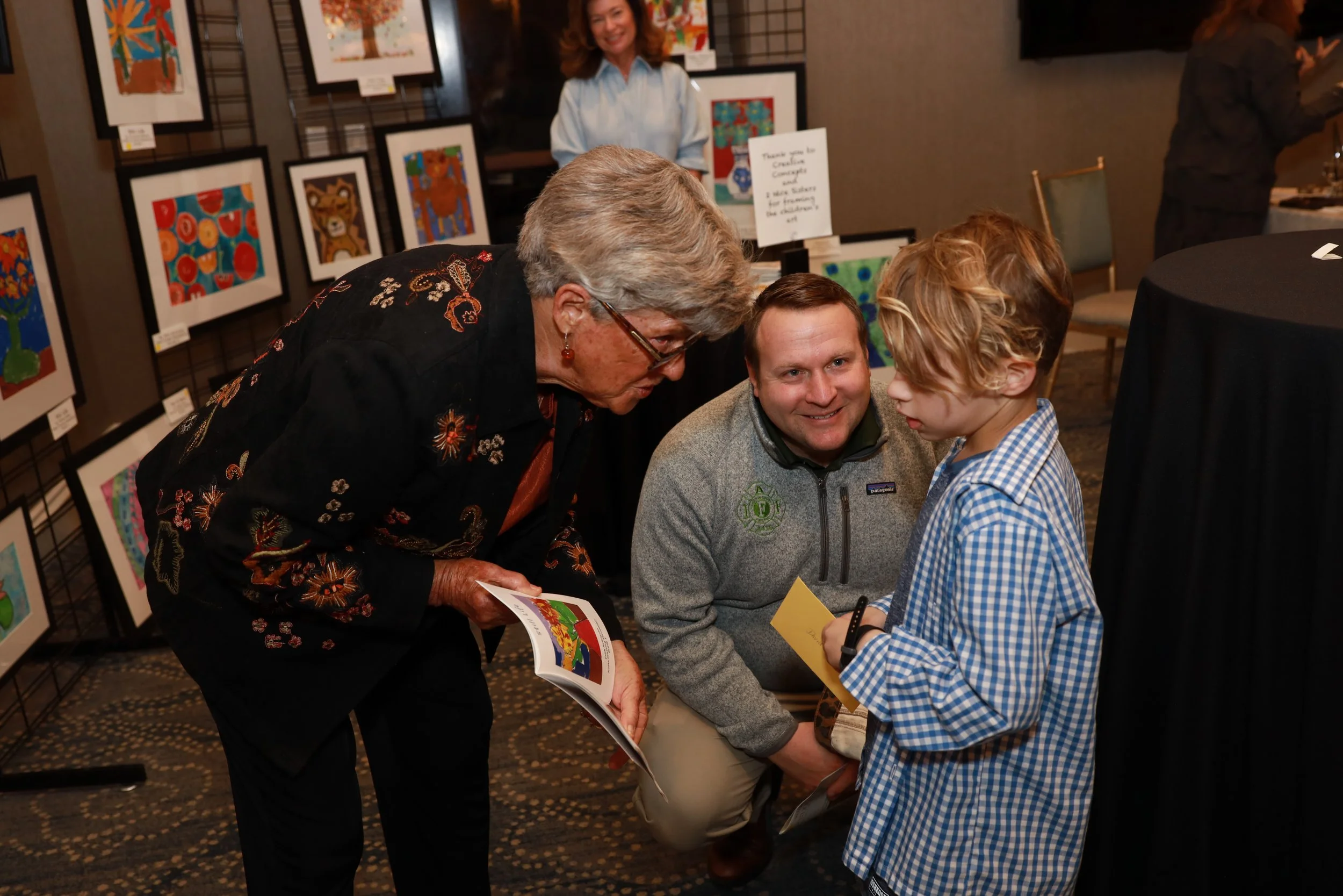 A group of four people, including an elderly woman, a young boy, a middle-aged man, and a woman in the background, at an art exhibit. The elderly woman is showing a book or brochure to the boy, who is holding a yellow card or paper. The background fe