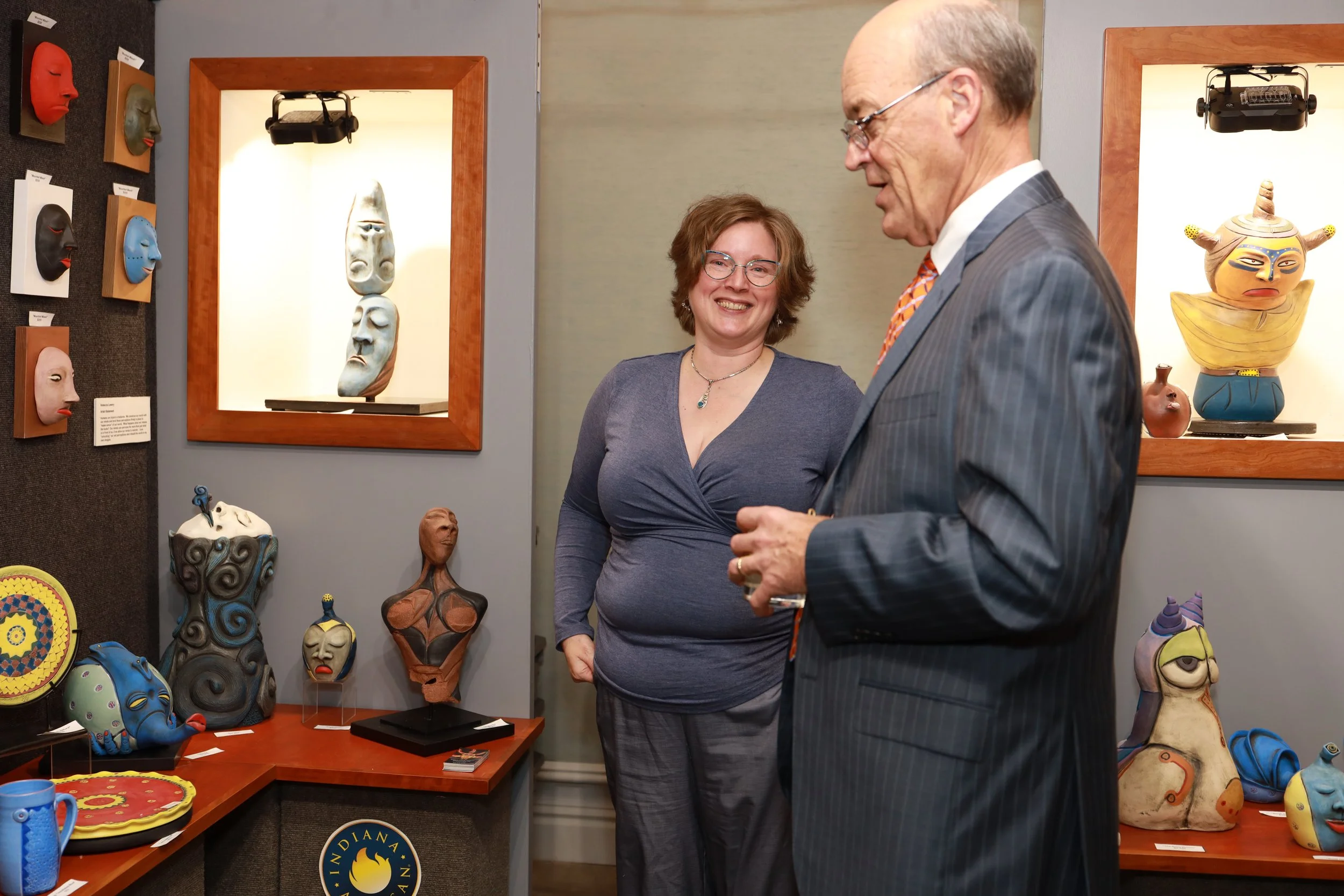 Two people, a woman with glasses and a man with glasses and a suit, are standing in front of a display of traditional African masks and sculptures in an art gallery or museum.