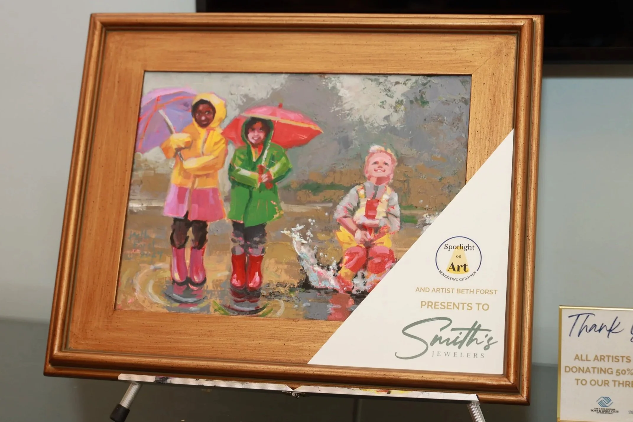 Painting of three children standing outdoors with umbrellas in rainy weather, dressed in colorful raincoats and rain boots, with a cloudy sky background.
