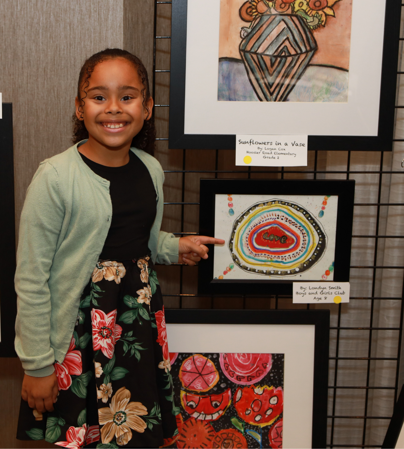 A young girl with curly hair and a big smile standing next to her artwork titled 'Sunflowers in a Vase' at an art exhibit. She is pointing at a colorful piece with concentric rings and the word 'Love' in the center, with several other paintings displayed behind her.