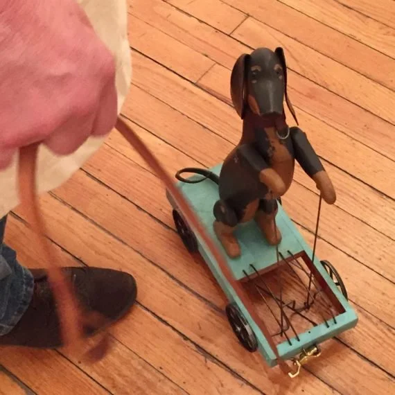 A wooden mechanical dog figure standing on a small blue skateboard with four black wheels, connected to a string held by a person, on a wooden floor.