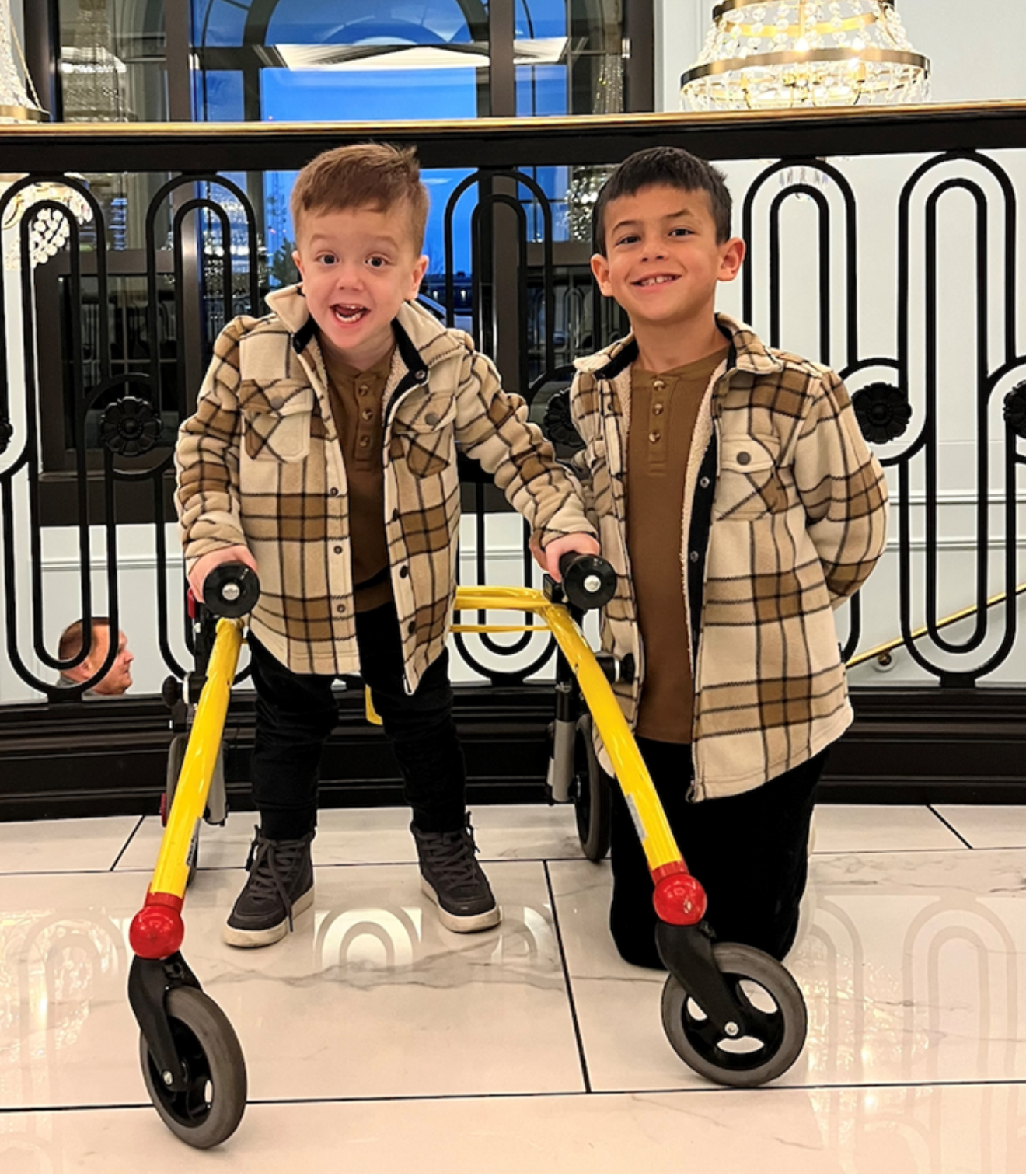 Two young boys wearing tan plaid jackets and brown shirts posing indoors. One boy is holding a yellow walker, smiling with his mouth open, while the other boy stands next to him, smiling. Decor and a balcony railing are visible in the background.