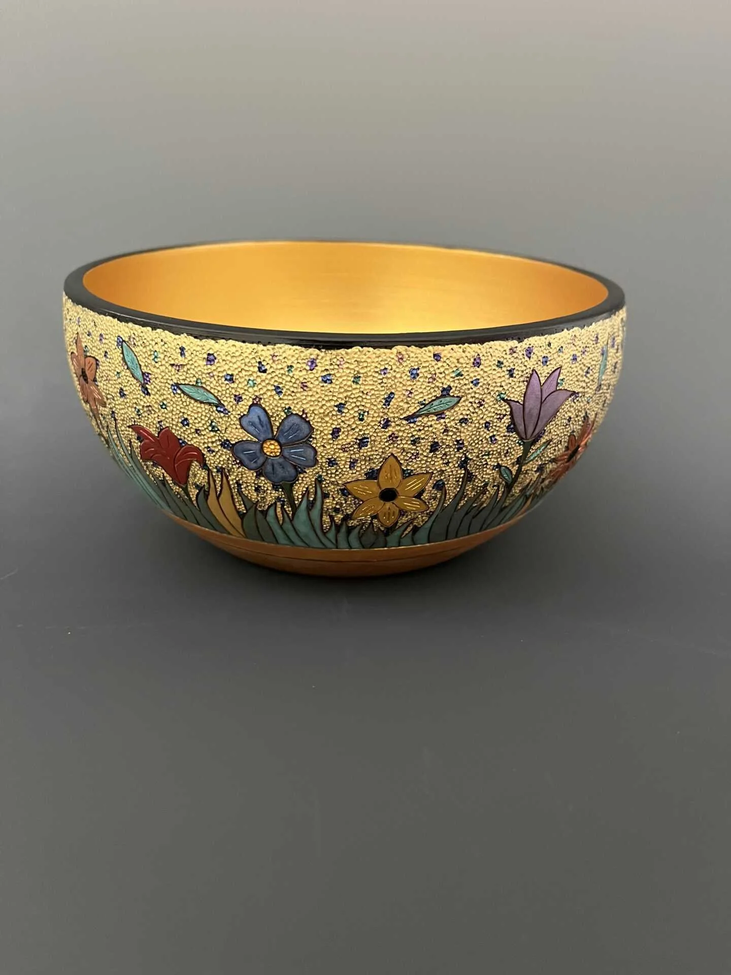 Decorative bowl with colorful floral design, gold interior, and black edge, set against a dark gray background.