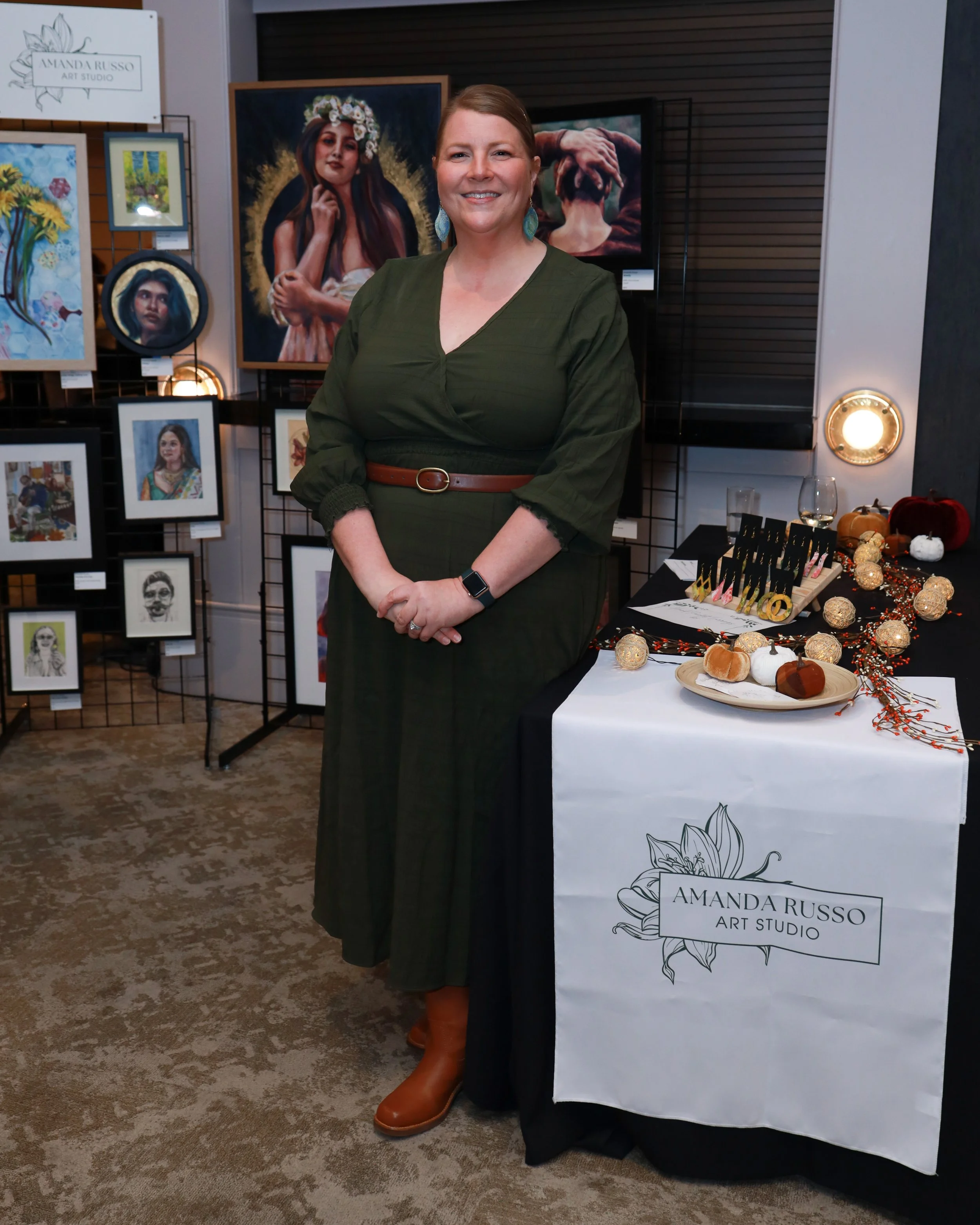 A woman with brown hair in a bun, wearing a dark green dress with a brown belt and brown boots, standing at an art exhibit. Behind her, there are various framed art pieces on display, including portraits and paintings of different styles. To her righ