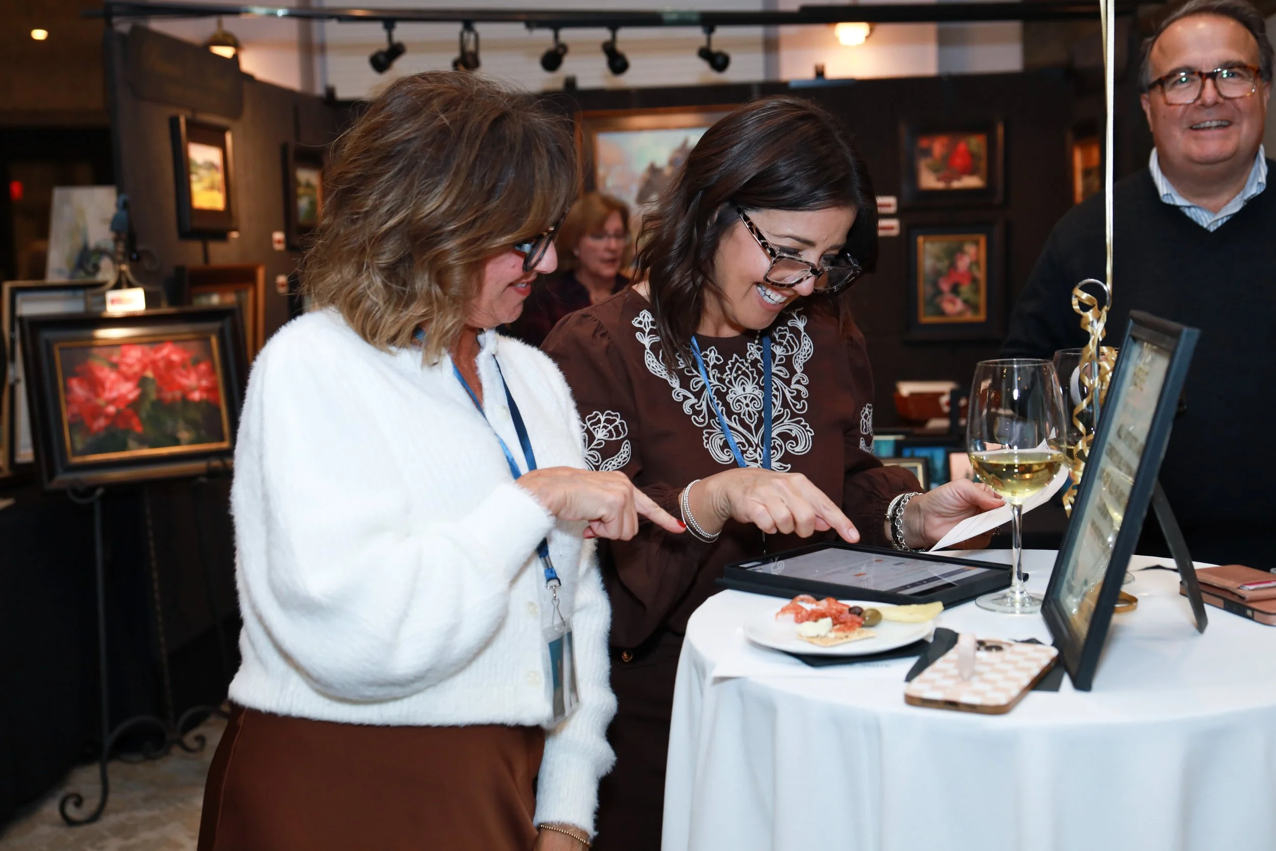 Three women and a man are at an art exhibit, looking at framed artwork on display. The women are smiling and appear to be enjoying themselves. One woman is holding a glass of white wine.