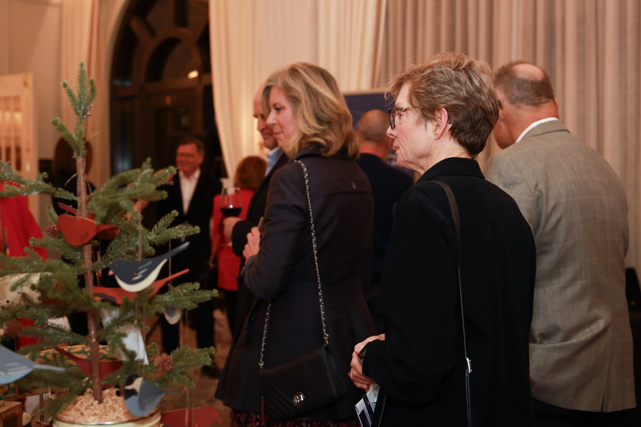 People gathered around a display table with Christmas decorations, including small pine trees, ornaments, and ribbons, in a warmly lit room with beige curtains in the background.