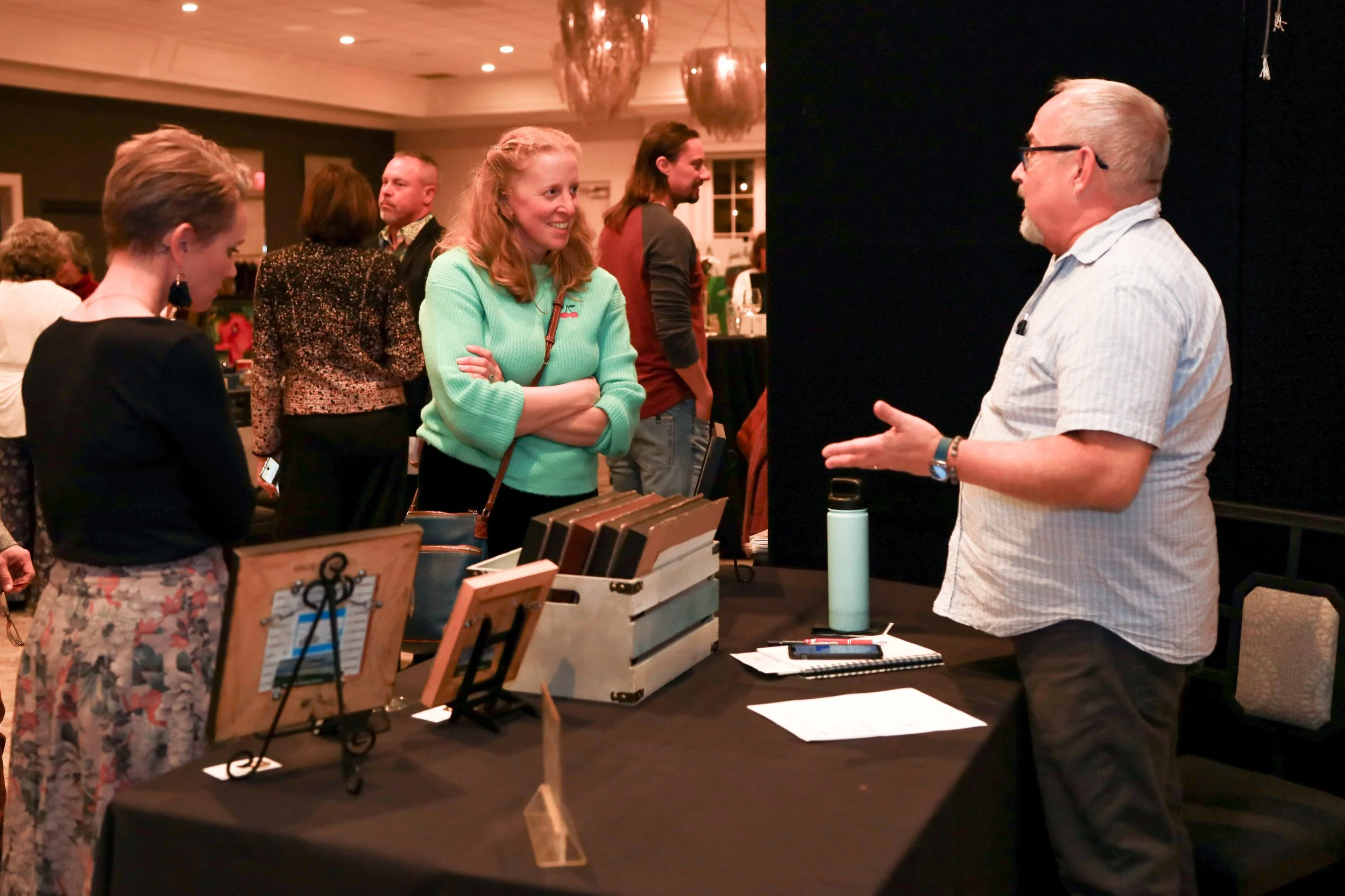 People standing and chatting at an indoor event, with tables displaying books and other items, some engaged in conversation, in a warmly lit room.