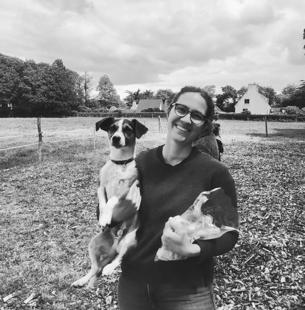Une femme souriante porte un chien de race indéfinie dans un champ. La femme porte des lunettes noir et un sweat sombre. Le chien a des oreilles droites et une expression attentive.