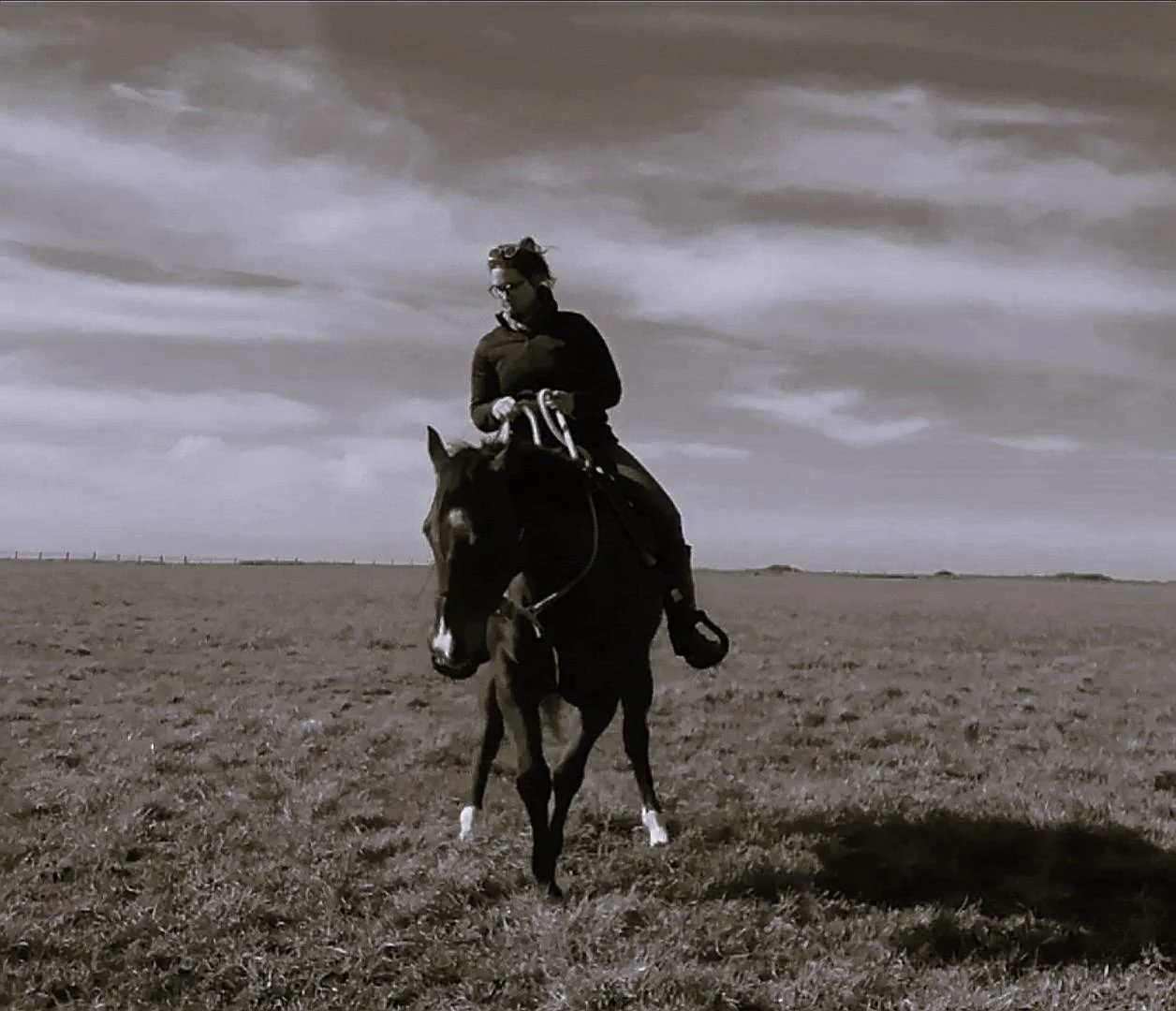 Une personne chevauche un cheval dans un champ ouvert sous un ciel nuageux en noir et blanc.