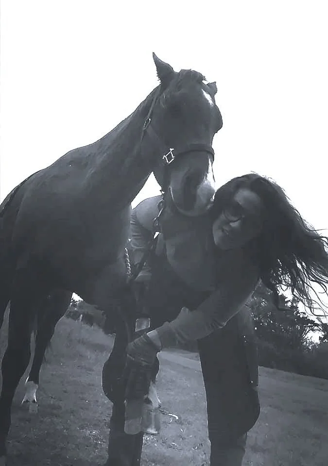 Une femme se tient à côté d'un cheval noir dans un champ en extérieur, dans un style noir et blanc.