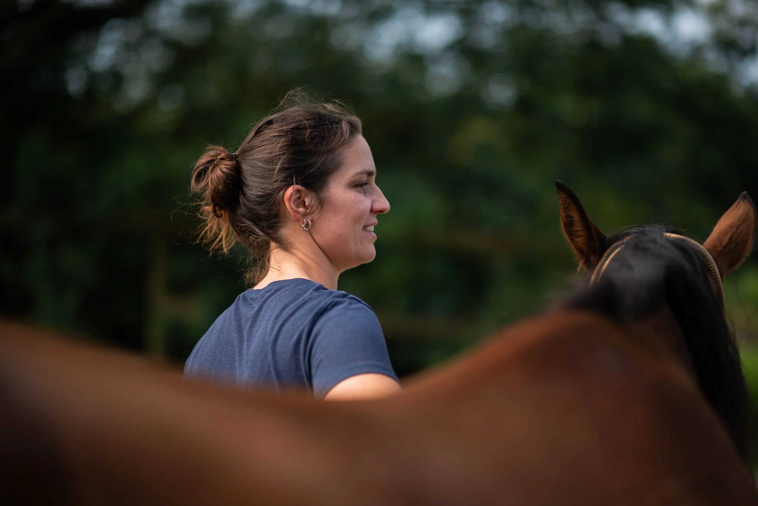 Une femme avec des cheveux attachés en chignon, souriante, portant un t-shirt bleu, est assise à côté d'un cheval brun dans un environnement naturel avec des arbres flous en arrière-plan.