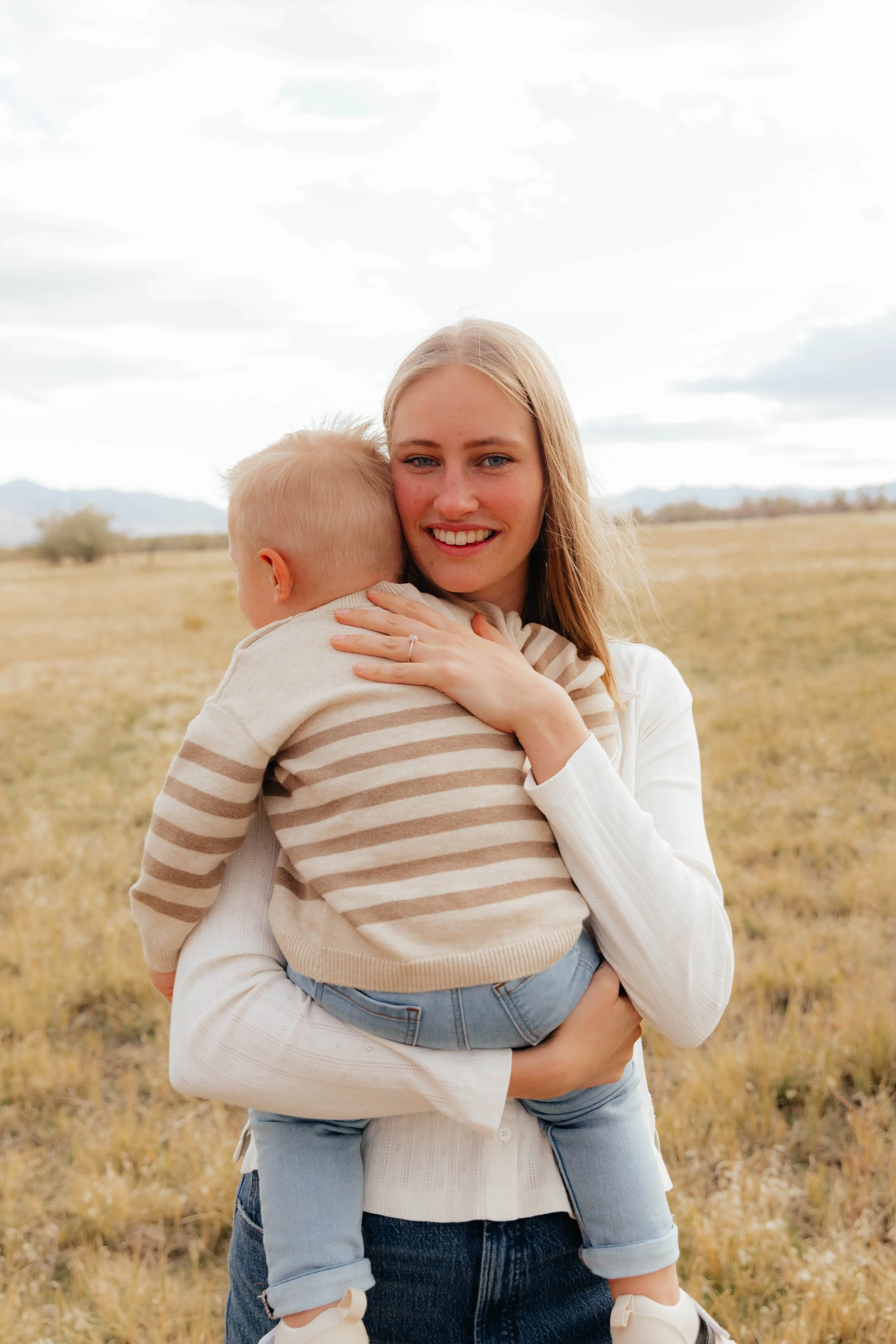 A woman smiling and holding a young child in a field with mountains in the background.
