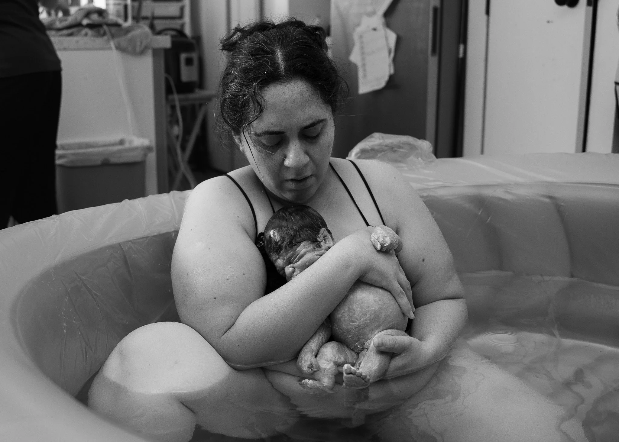 A woman in black and white holds a newborn baby in a birthing room, with medical equipment and household items in the background.