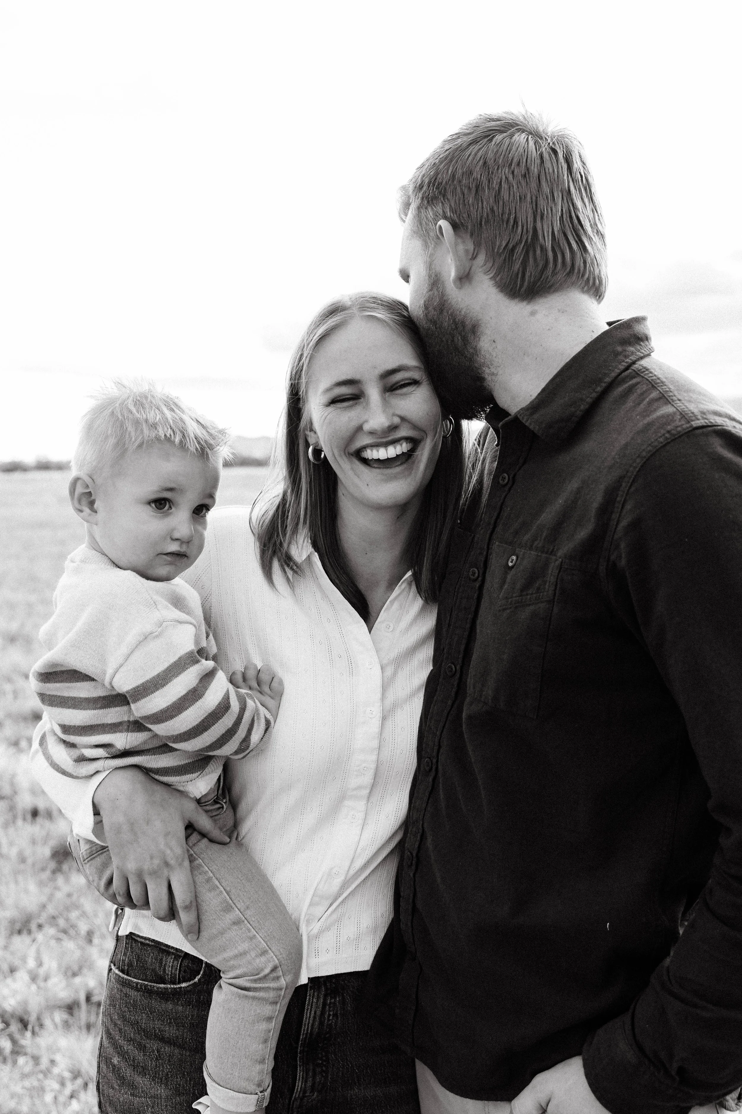 A black and white photo of a smiling woman holding a young boy on her hip, and a man leaning in to give her a kiss on the forehead, outdoors in a field.