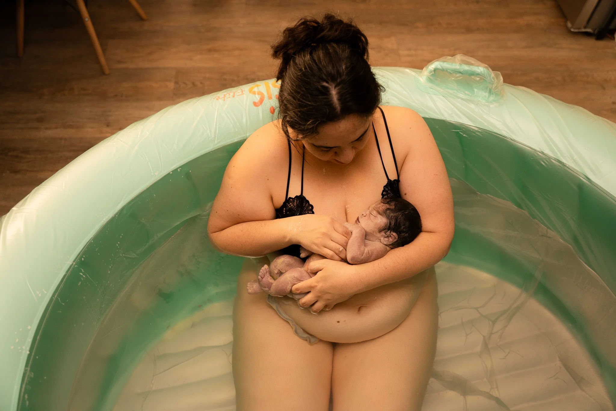 A woman with dark hair and a black top is sitting in a small inflatable pool, holding her newborn baby with dark curly hair. The woman is looking down at the baby, who is cradled in her arms.