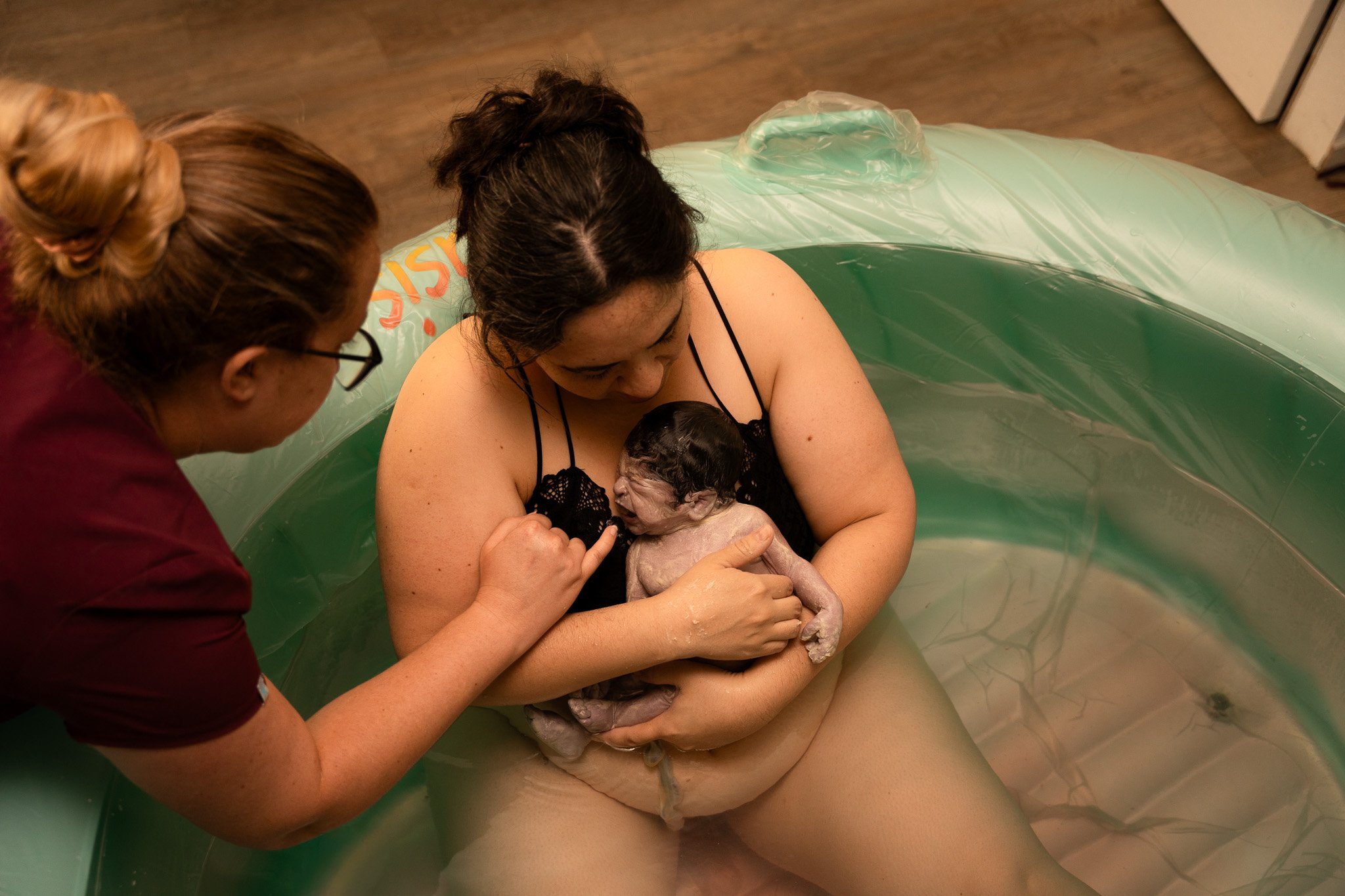 A woman holding her newborn baby in her arms during a water birth, assisted by a woman with glasses in a room with wooden flooring.