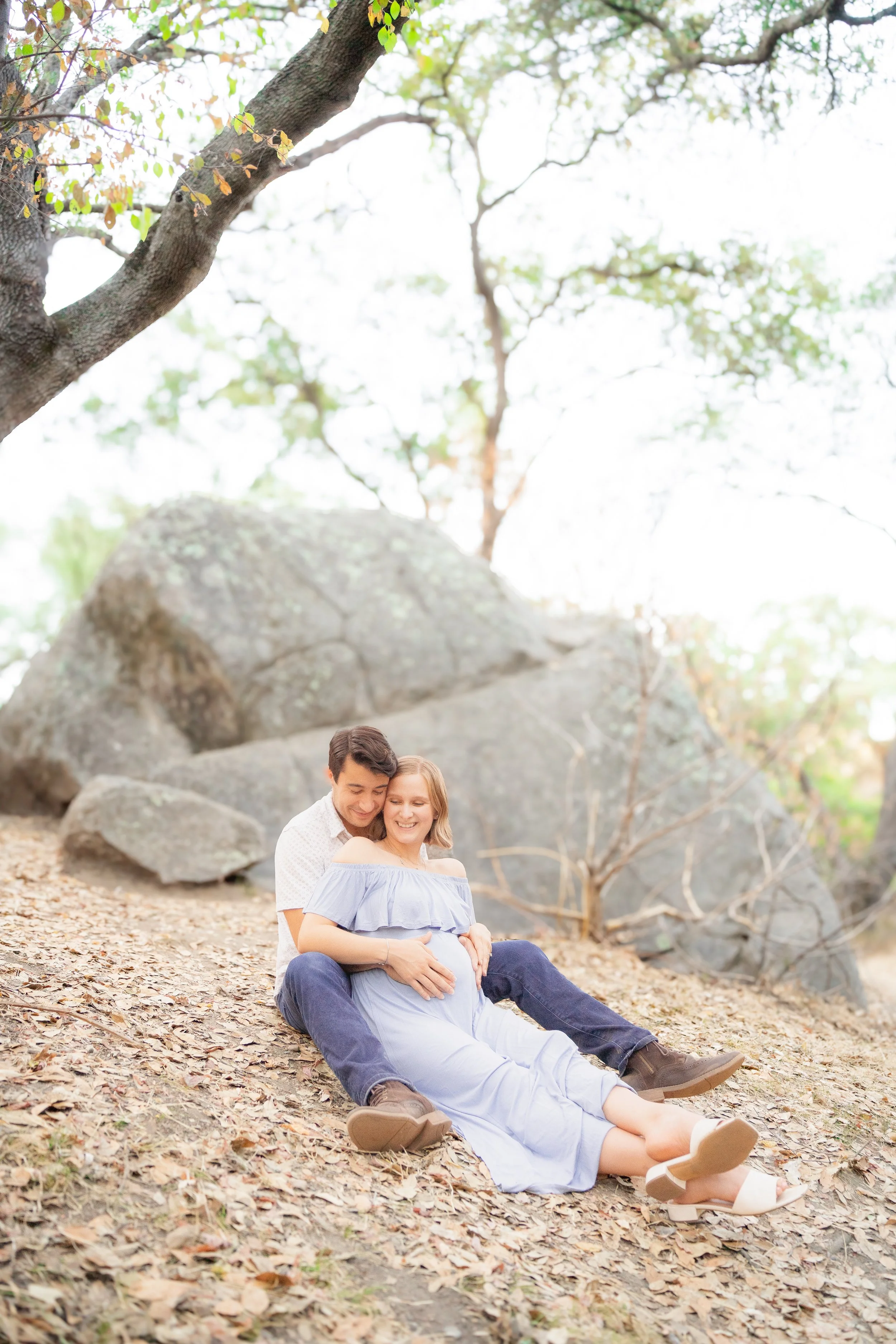 A man and woman sitting on the ground in a wooded area, with the woman appearing pregnant. The man is holding the woman from behind, and they are both smiling. There are large rocks and trees in the background, with fallen leaves on the ground.