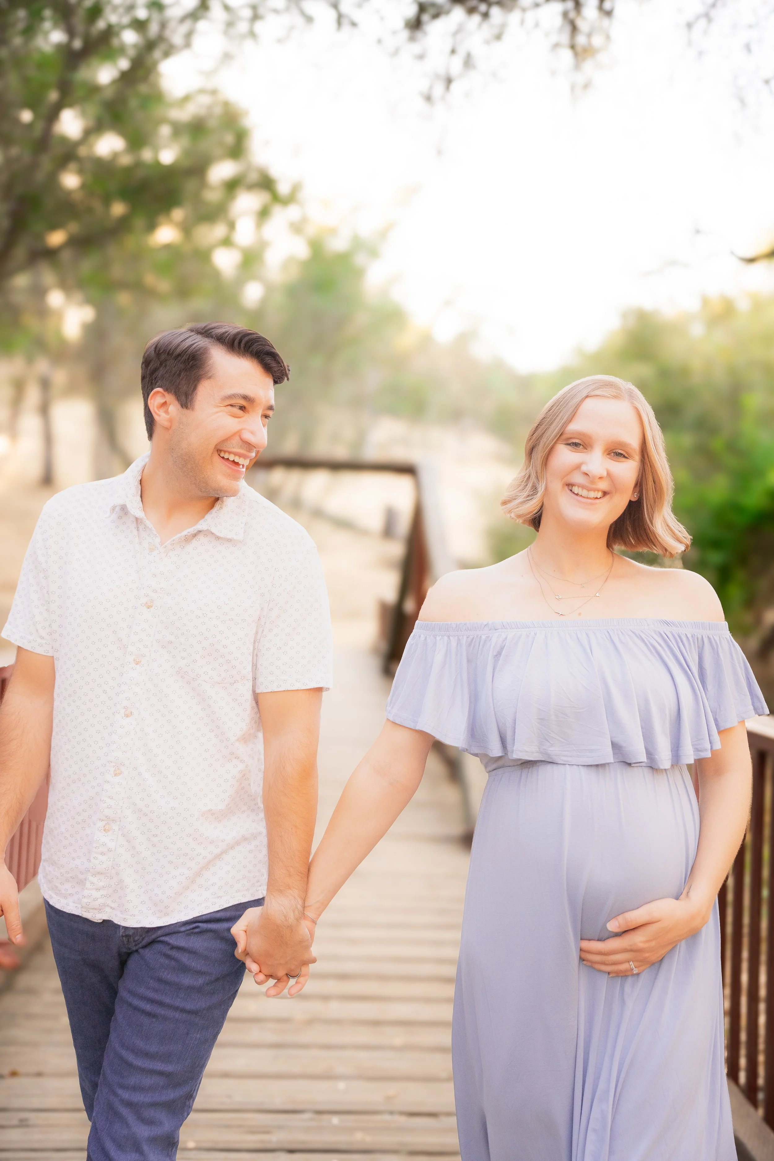 A smiling couple holding hands and walking outdoors on a wooden pathway, with trees and a bench in the background, during a sunny day.