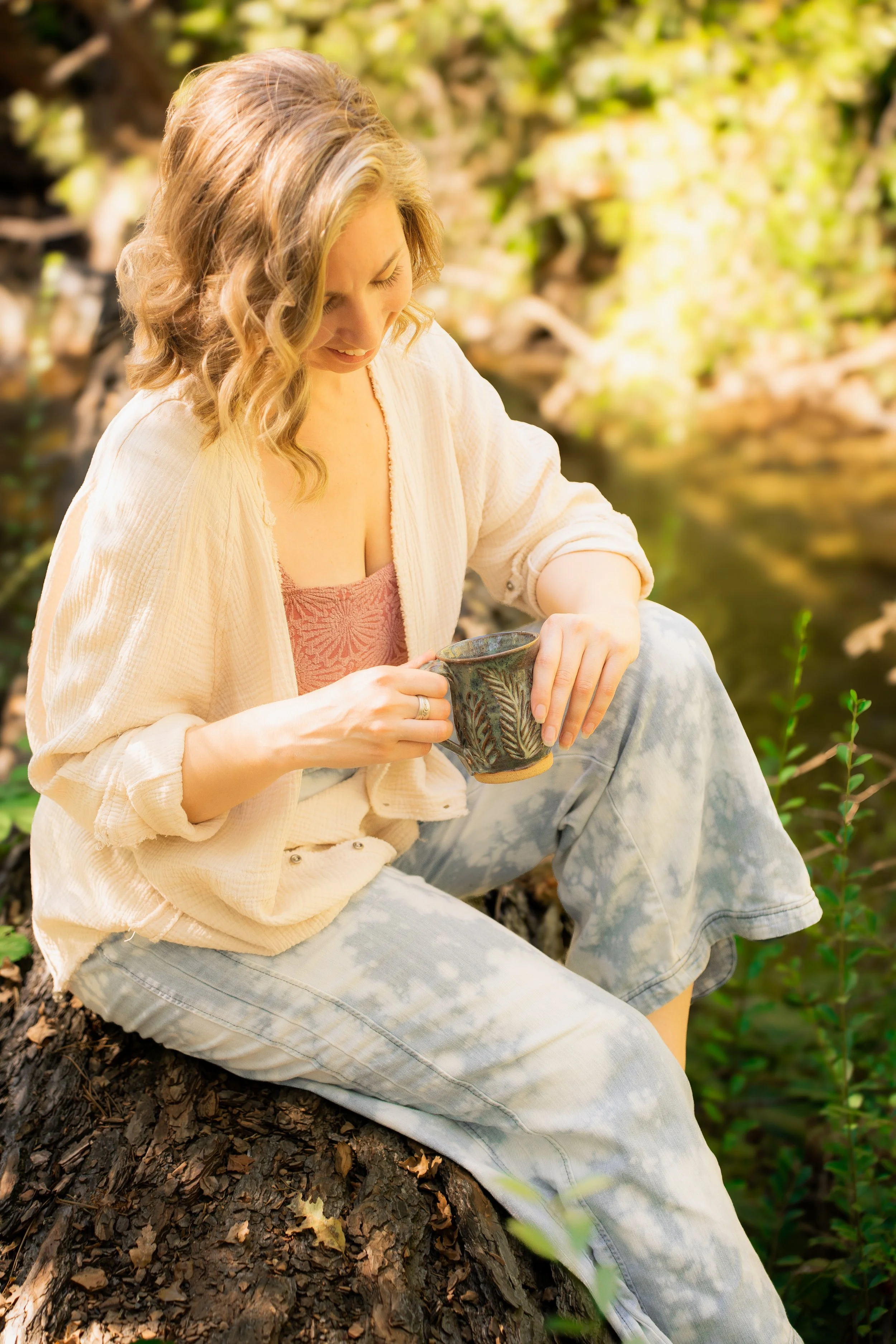 A woman with wavy blonde hair sitting on a tree root beside a creek, holding a mug and looking down with a gentle smile.
