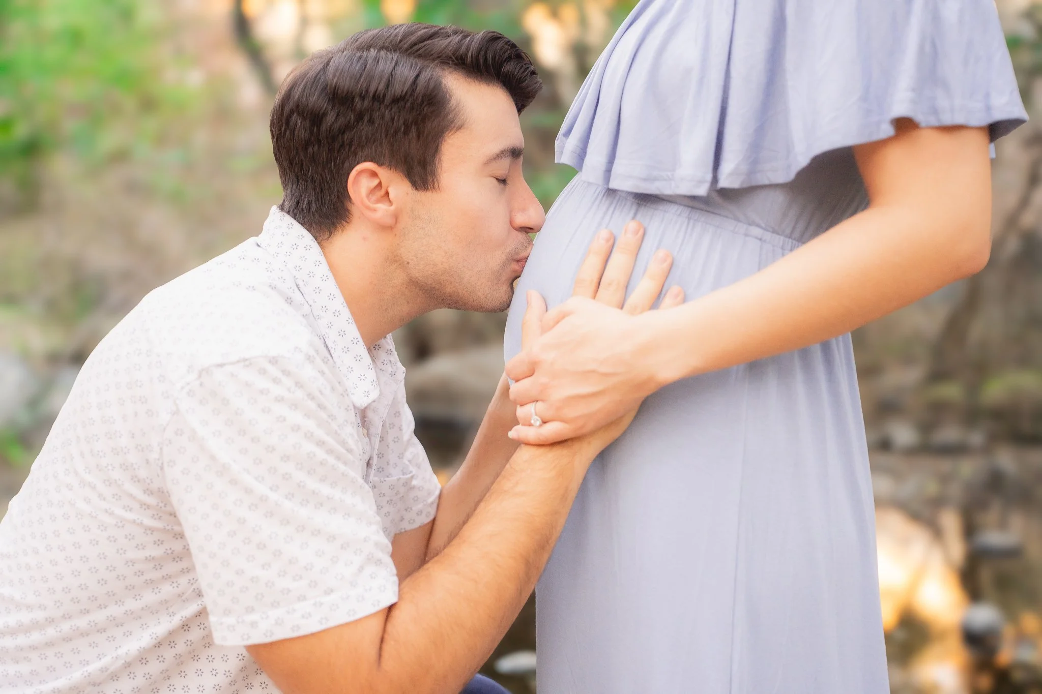 A man kissing a pregnant woman's belly outdoors.