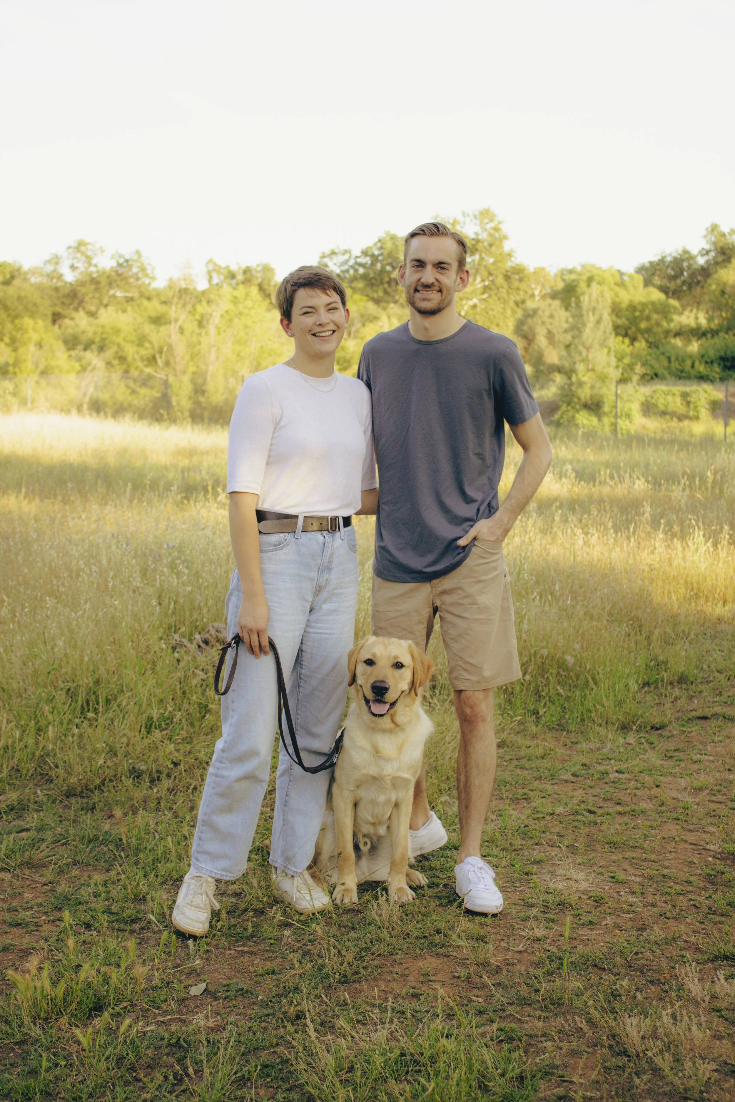 A smiling woman and man standing outdoors in a grassy field with greenery and trees in the background, holding a leash attached to a happy golden retriever dog.