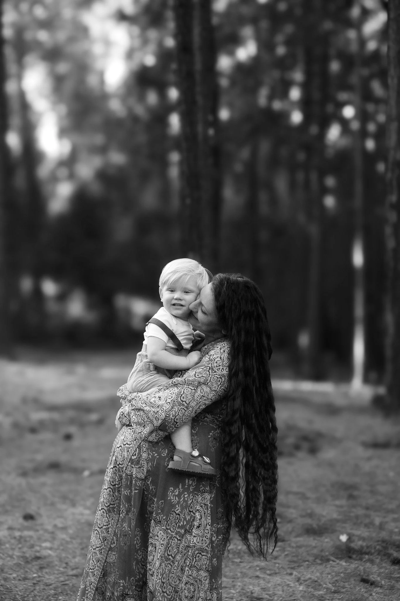 A woman with long dark hair holding a young boy with short light hair outdoors in a park, blurred trees in the background in black and white.
