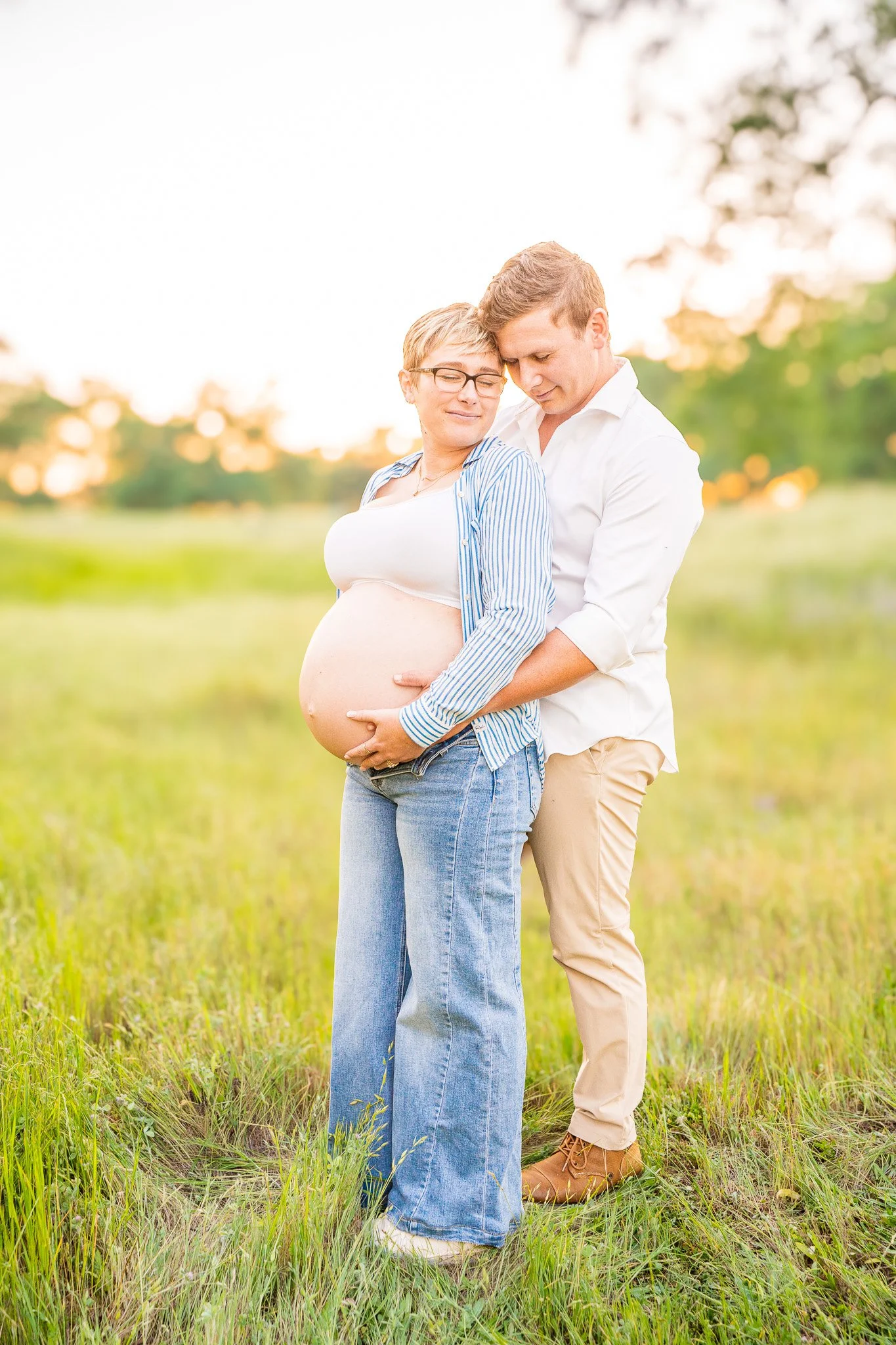 A pregnant woman and a man standing in a grassy field, embracing each other with their eyes closed, in warm sunlight with trees in the background.