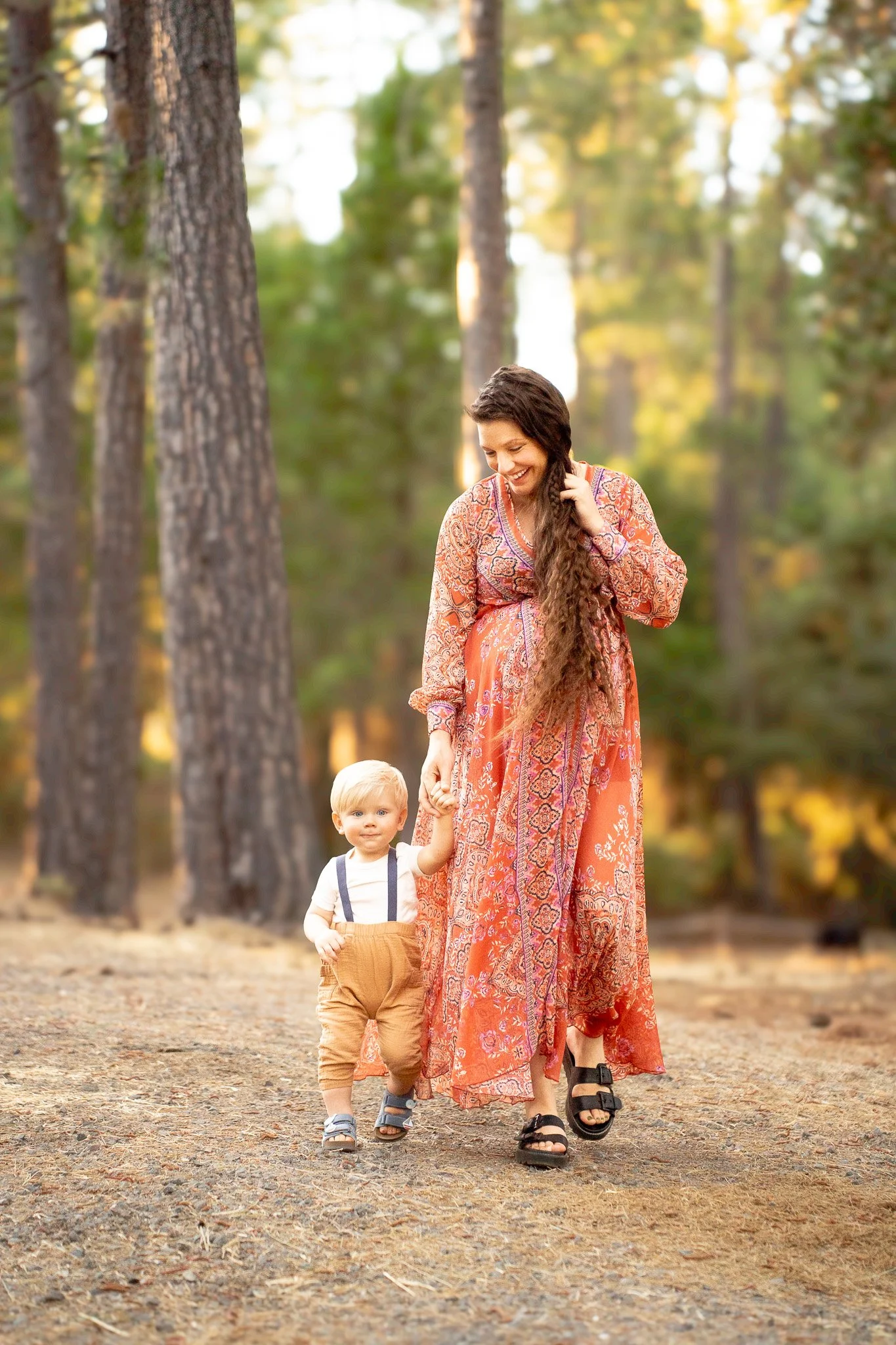 A woman with long brown hair smiling and holding hands with a young blonde-haired boy walking in a forest during autumn.