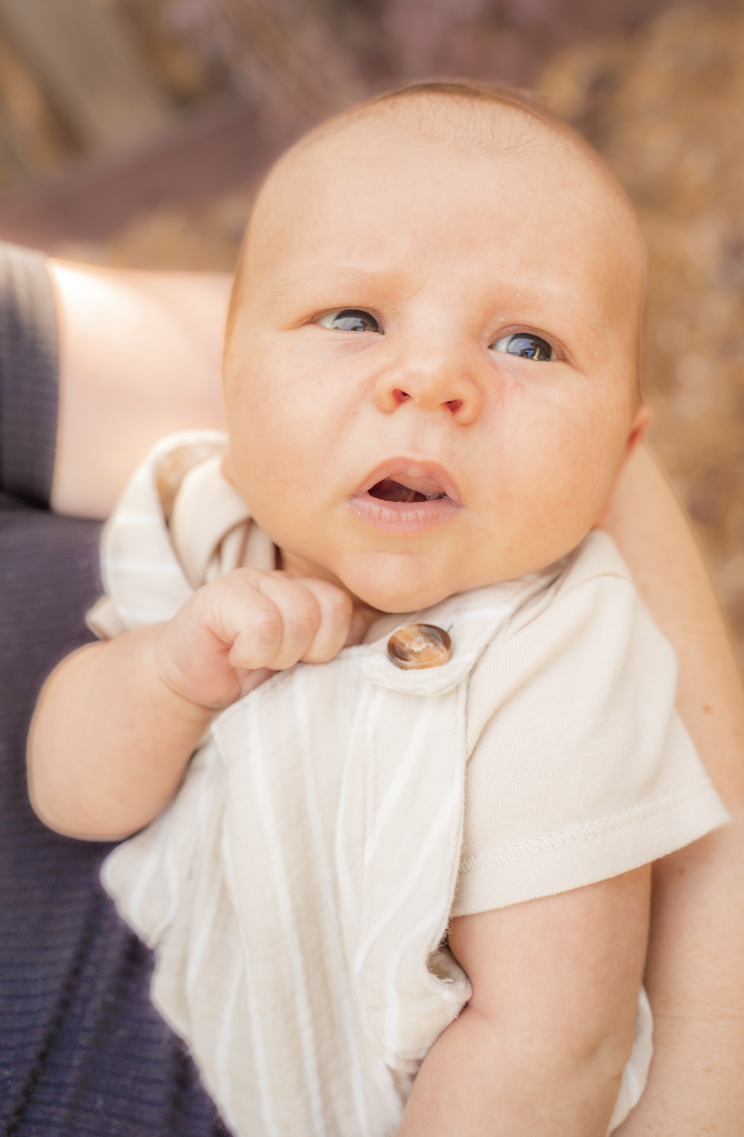 Close-up of a baby with light skin, blue eyes, and a slightly open mouth, being held and looking to the side outdoors.