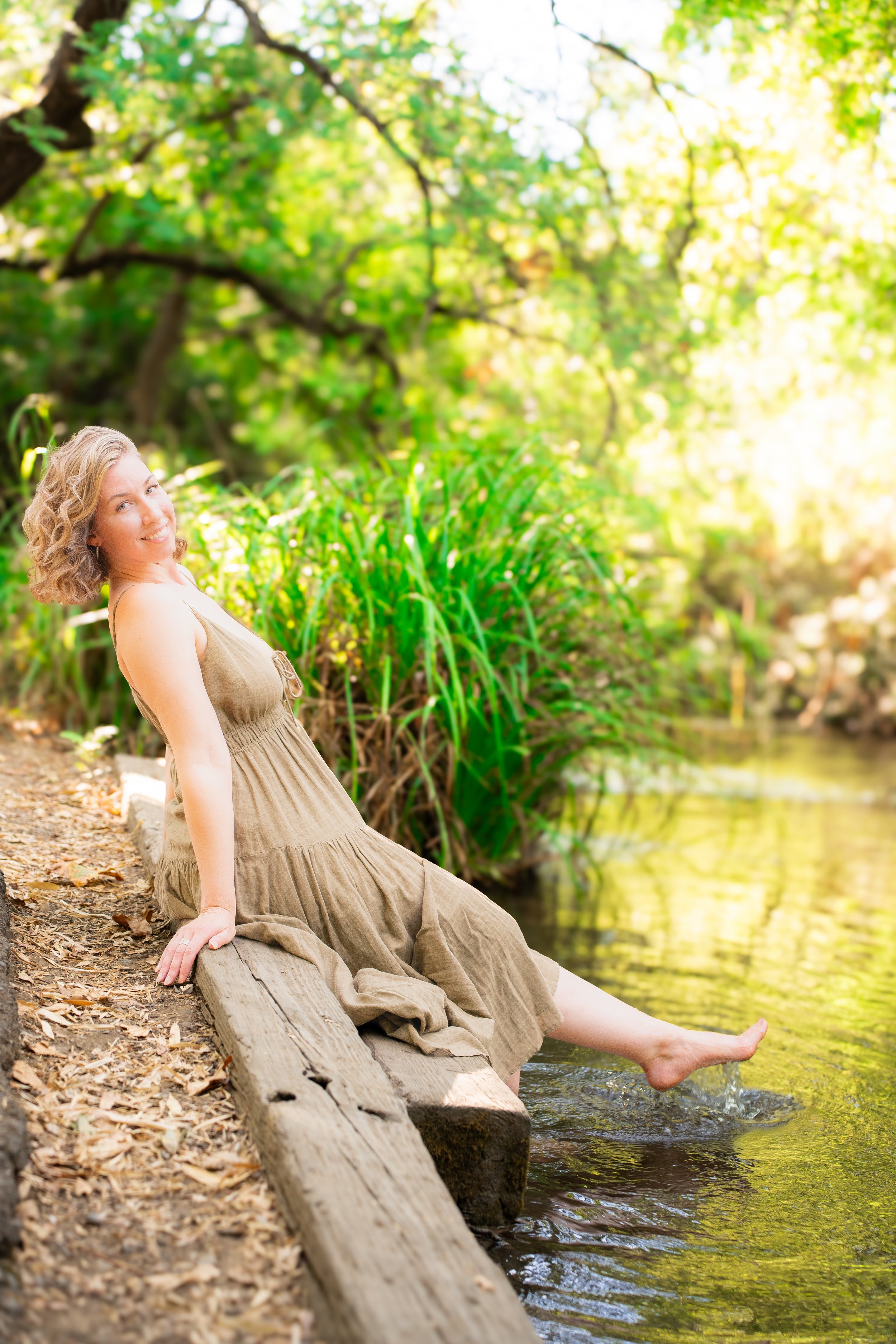 A woman with curly blonde hair sitting on a wooden dock beside a river, dipping her toes in the water, surrounded by lush green trees and foliage during daytime.
