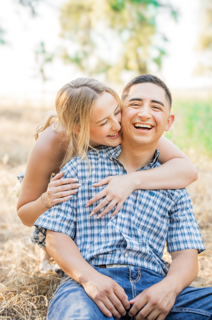 A young couple sitting outdoors, smiling and laughing, with the woman hugging the man from behind during daytime.