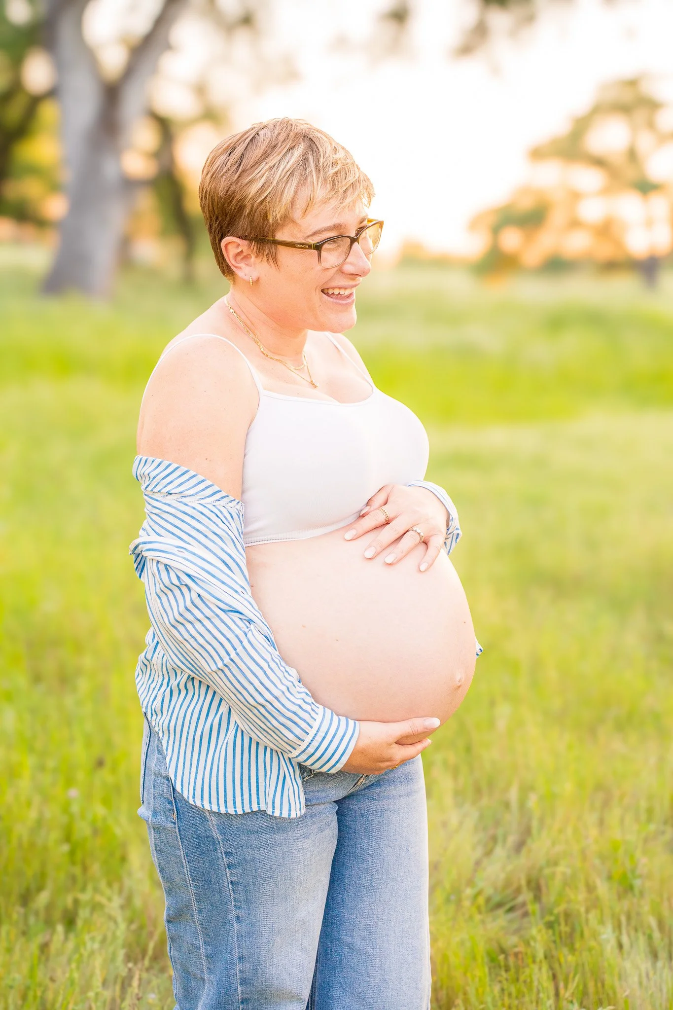 A pregnant woman standing in a grassy field with trees in the background, smiling and wearing glasses, a white tank top, and a striped shirt falling off her shoulders.