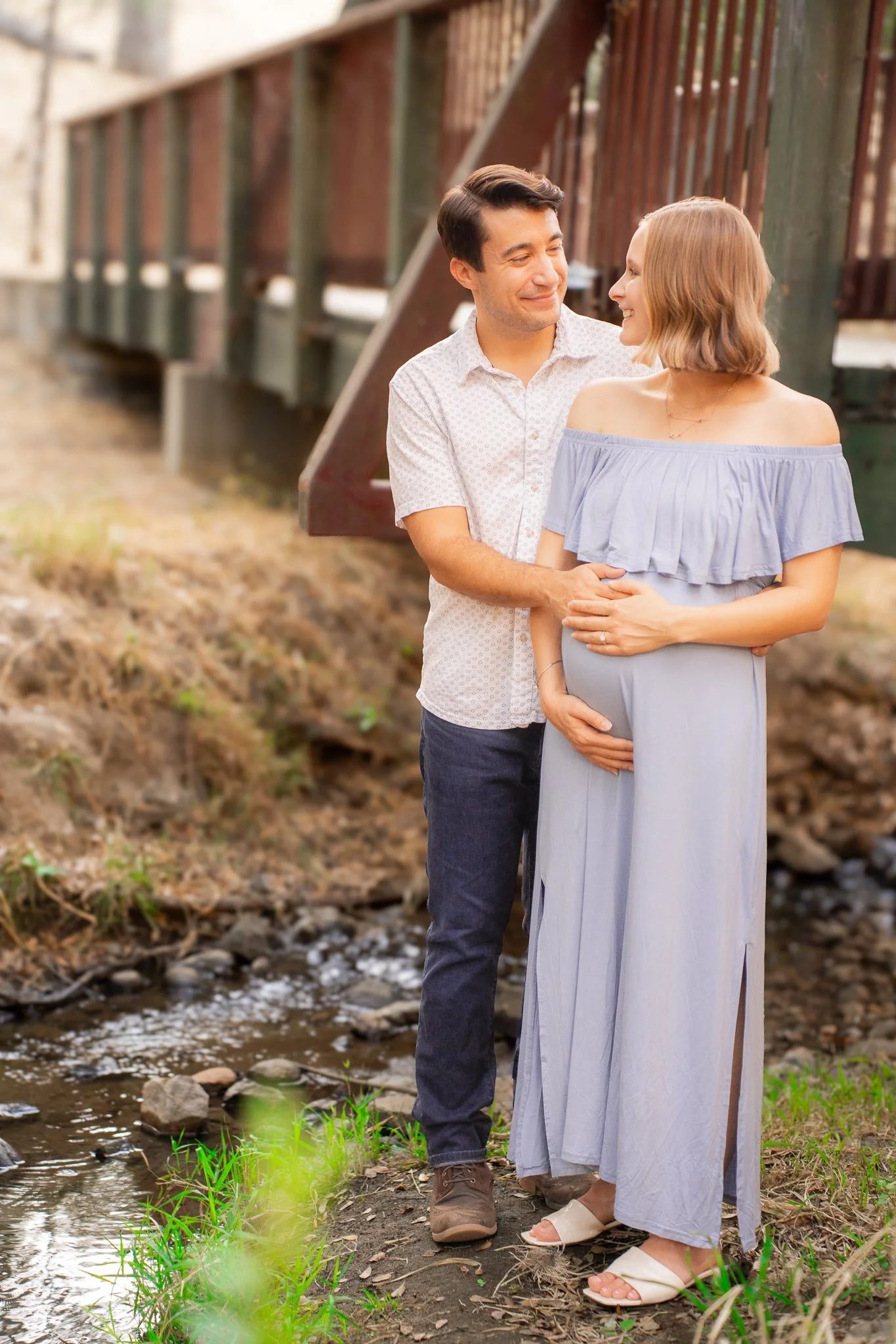 A couple standing close together by a small creek in a wooded area, smiling and looking at each other.