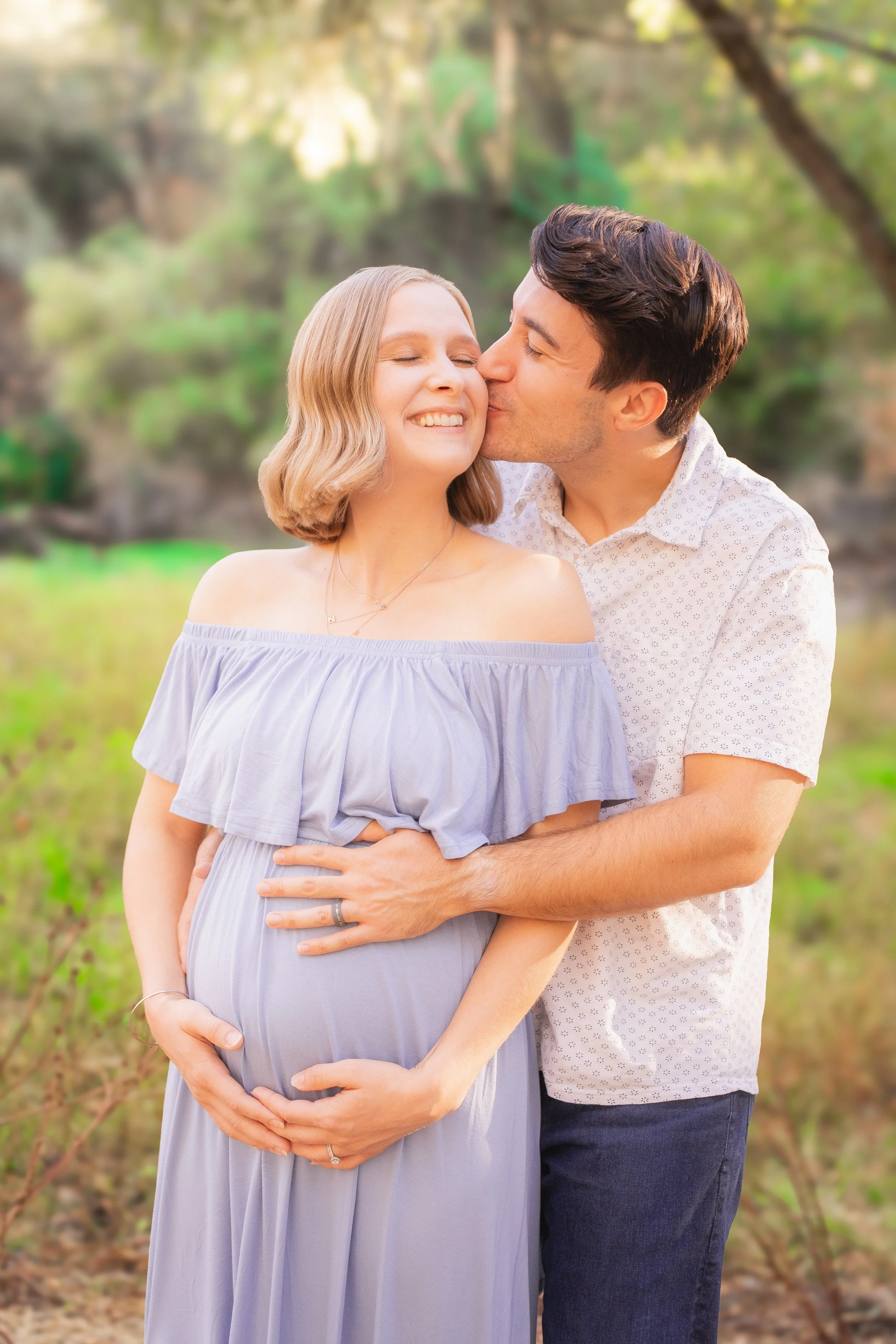 A couple standing outdoors, with the man kissing a pregnant woman's cheek as she smiles happily and cradles her baby bump.