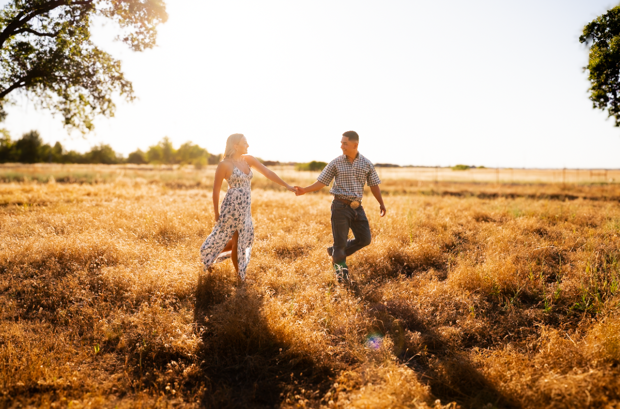 A couple holding hands and dancing in a sunlit open field during sunset, with trees in the background.