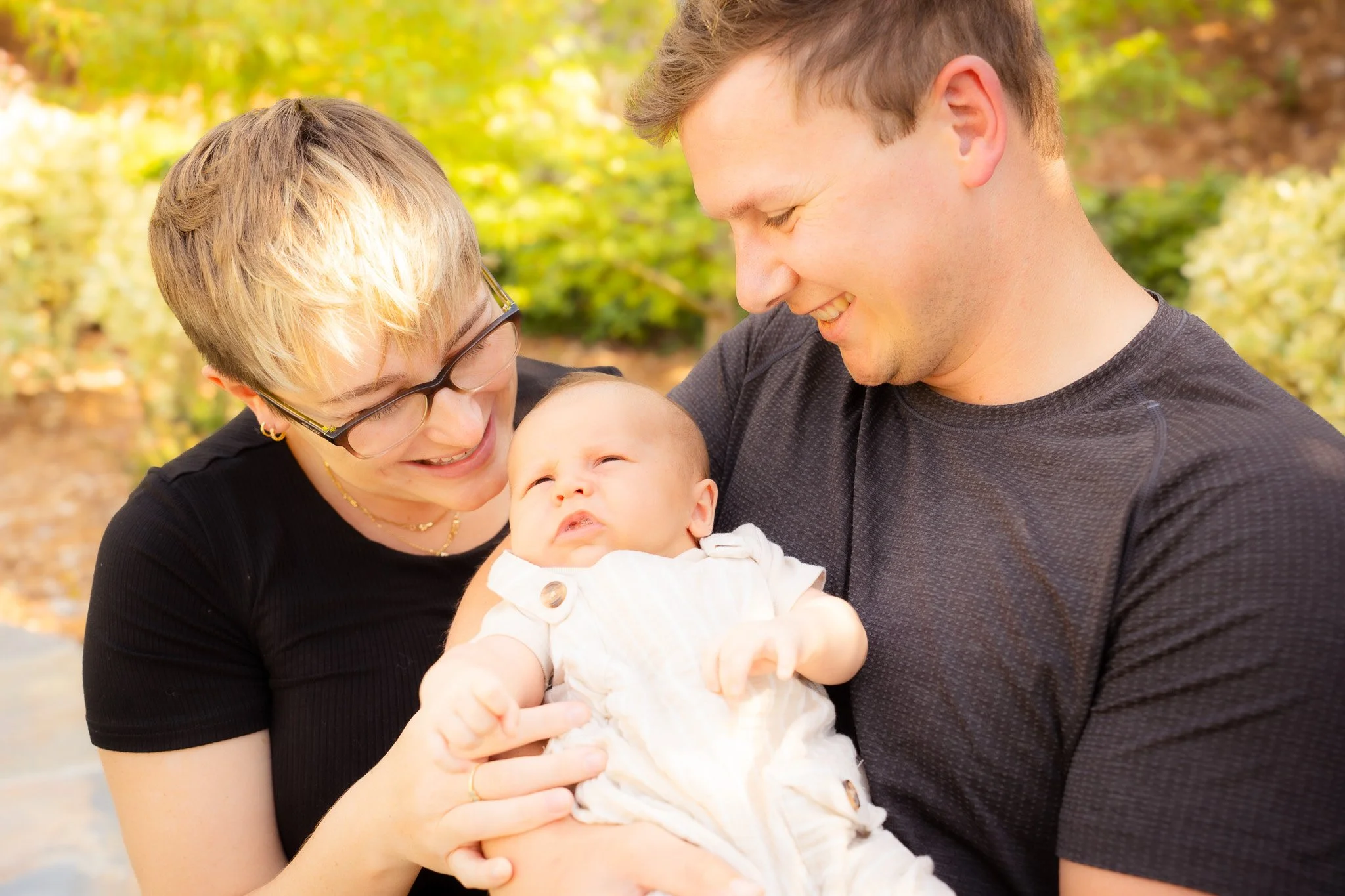 A smiling woman and man holding a baby outdoors with green and yellow foliage in the background.