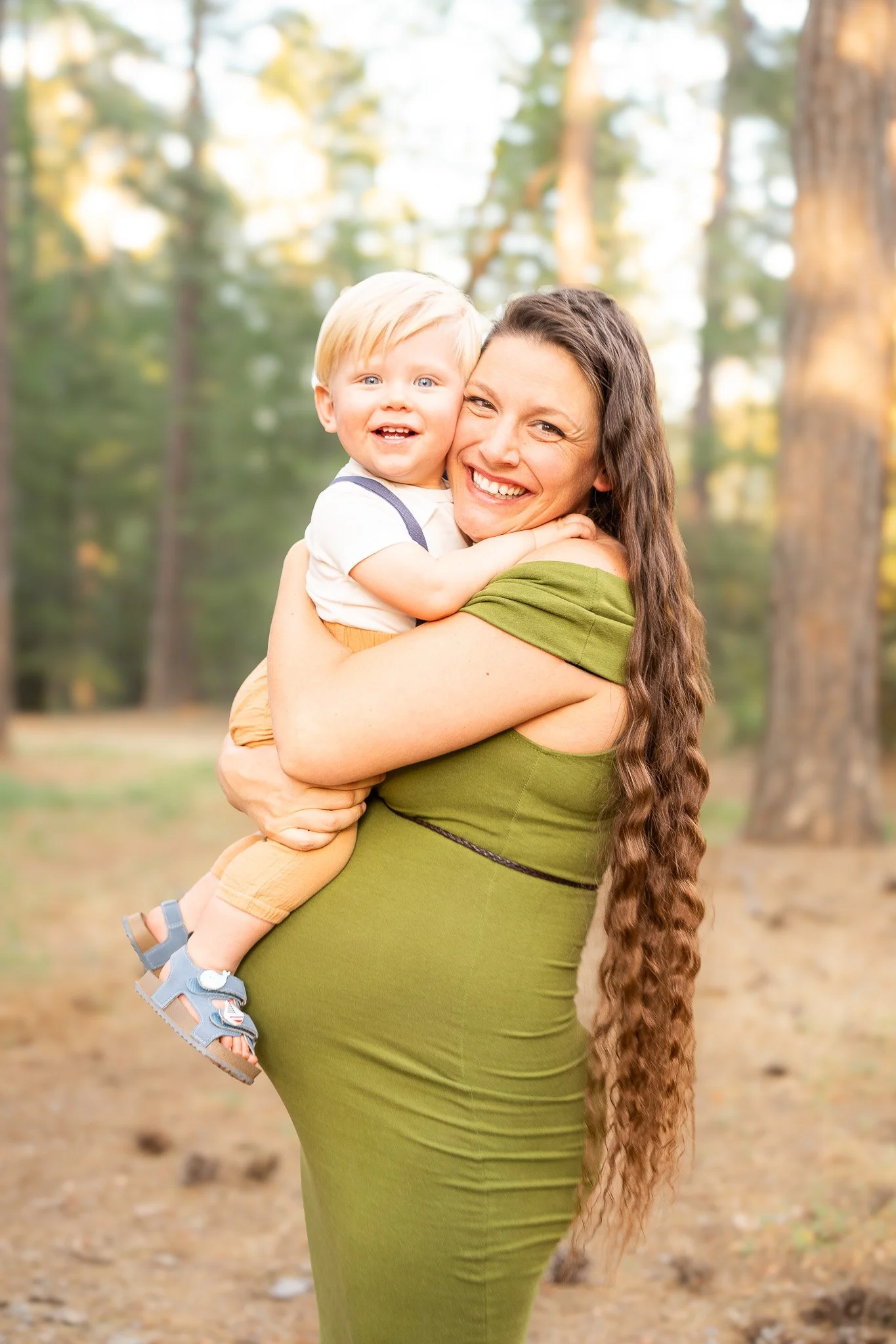 A woman with long, wavy brown hair wearing a green dress is smiling while holding a young blond-haired boy in her arms. They are outdoors in a forest setting with trees and a dirt path in the background.