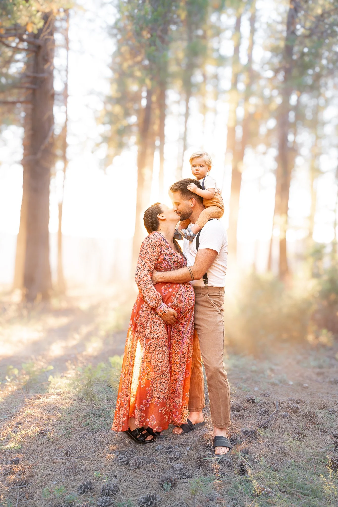 A family of three, a woman, a man, and a young boy, is standing outdoors in a forest with tall trees and sunlight filtering through the leaves. The woman and man are kissing, and the boy is sitting on the man's shoulders, looking at the camera.