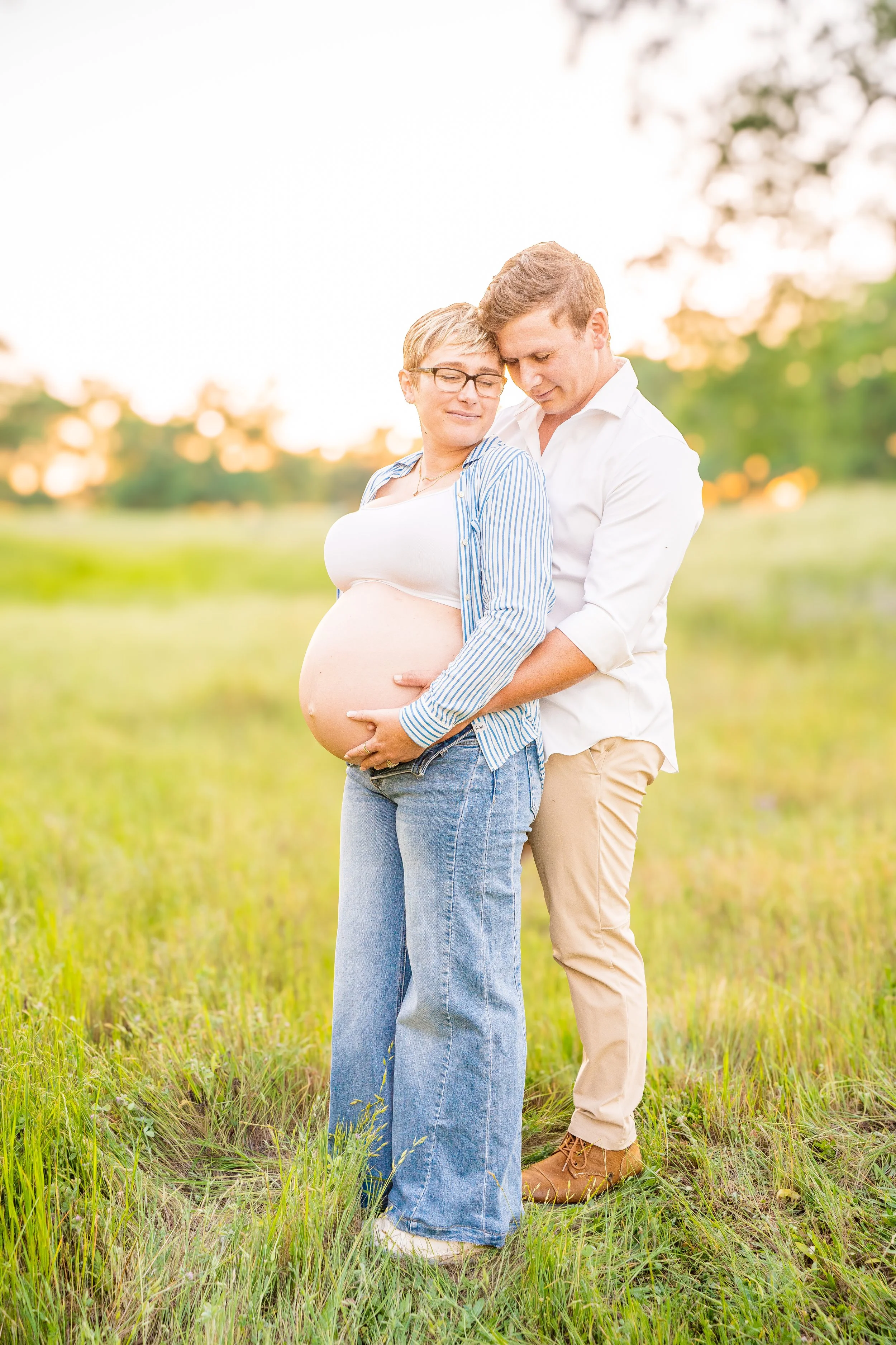 A pregnant woman with glasses and short hair in a striped shirt stands in a grassy field, cradling her belly, with a man in a white shirt and beige pants standing behind her, holding her and looking down affectionately at her belly, both in a warm embrace during sunset.