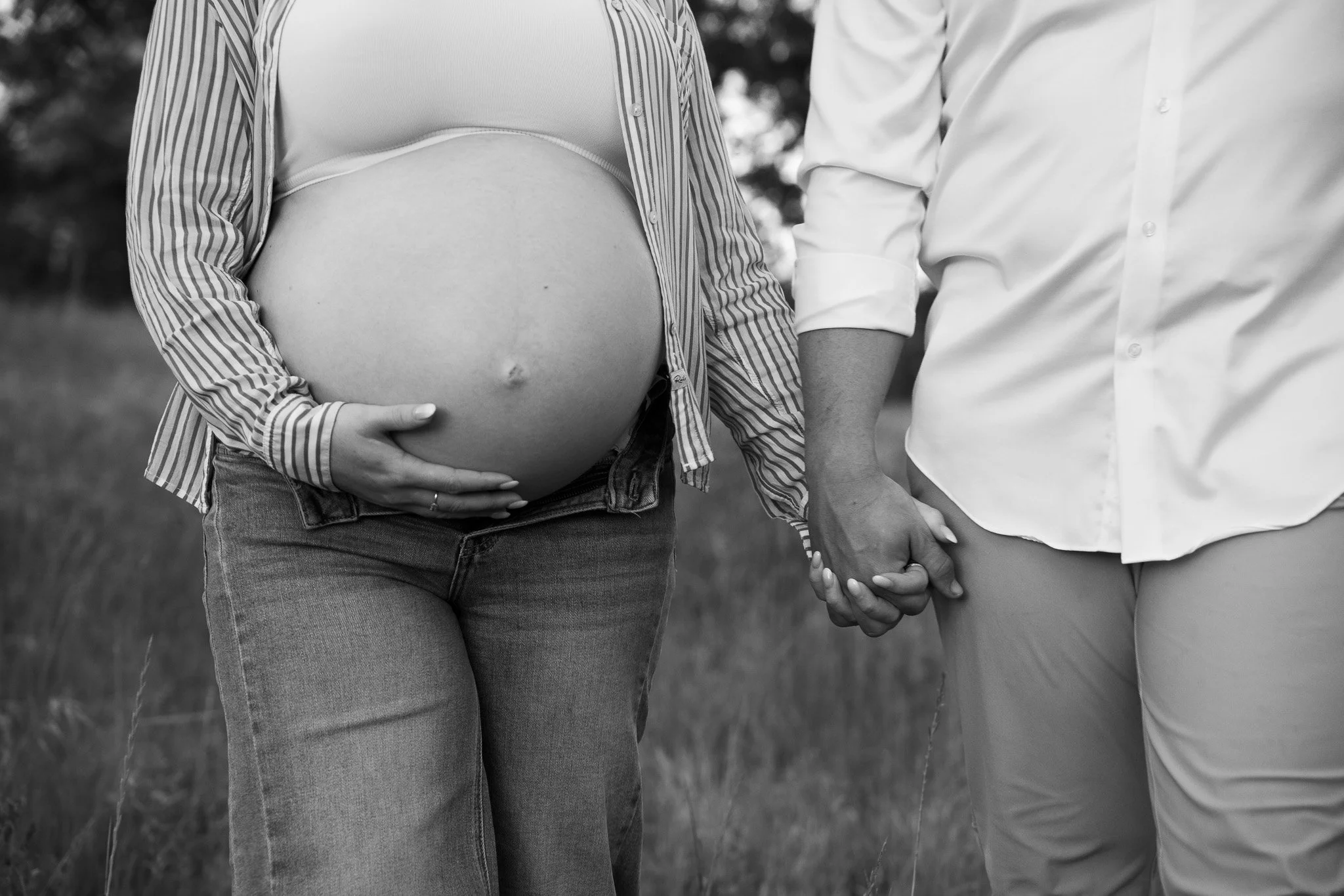 A pregnant woman wearing a striped shirt and jeans holds her belly, standing next to a man in a white shirt, with their hands joined, outdoors with blurred trees in the background.